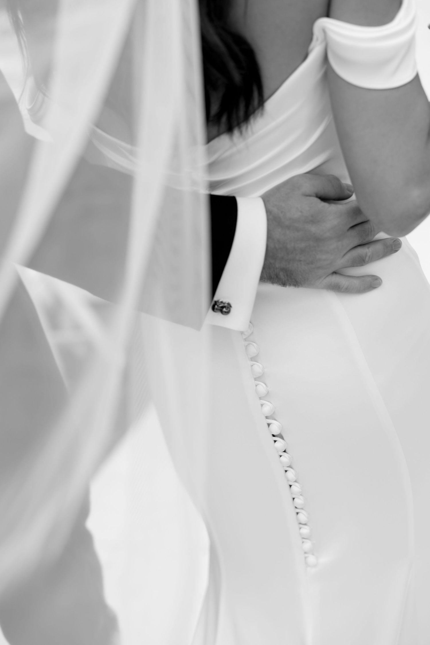 Black and white wedding photo showing bride and groom embracing, featuring elegant button-back dress and flowing veil.