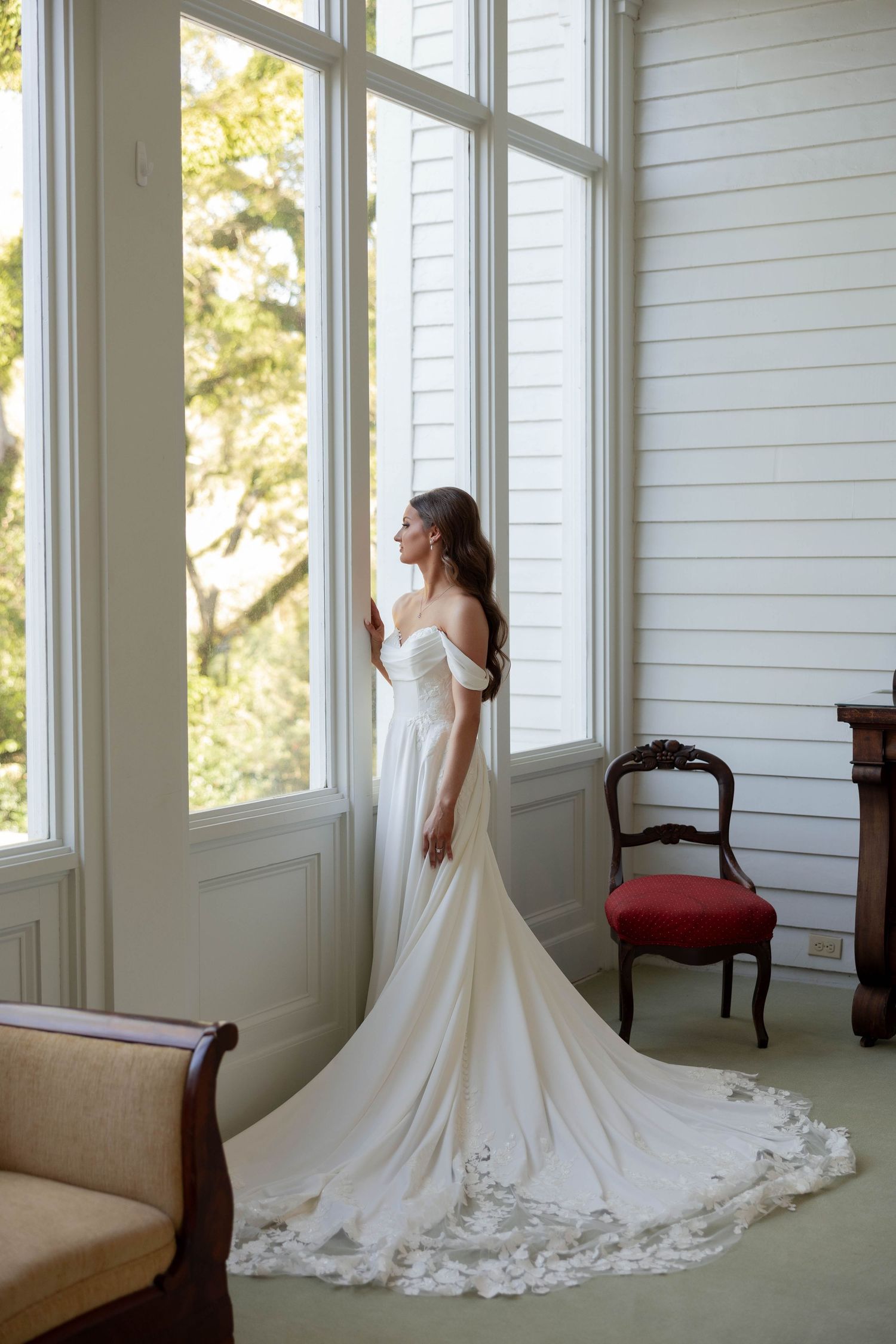 Elegant bride in off-shoulder wedding gown with lace train standing by tall windows in a sunlit vintage room.