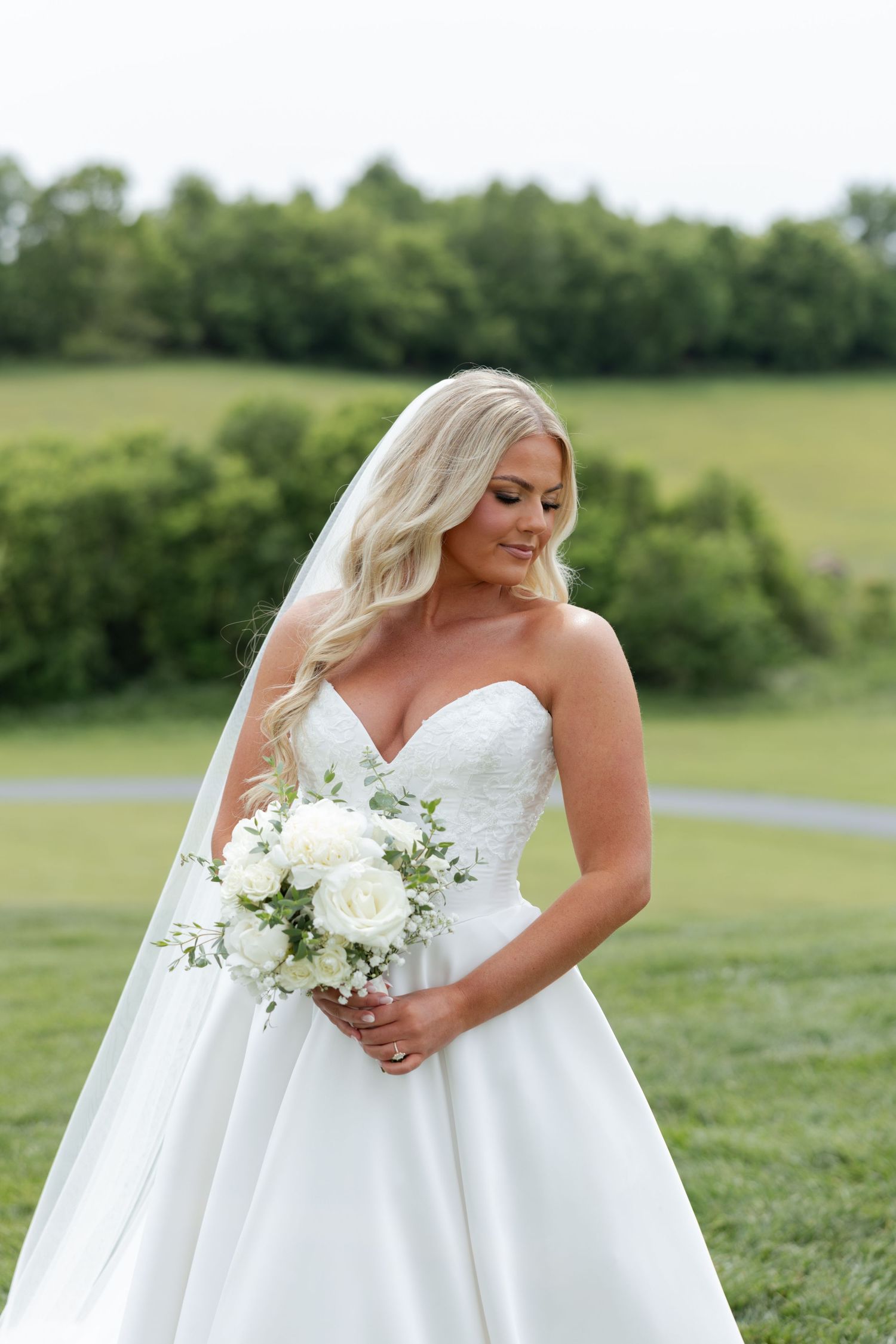 Blonde bride in strapless lace ball gown holding white rose bouquet in a lush green outdoor setting.