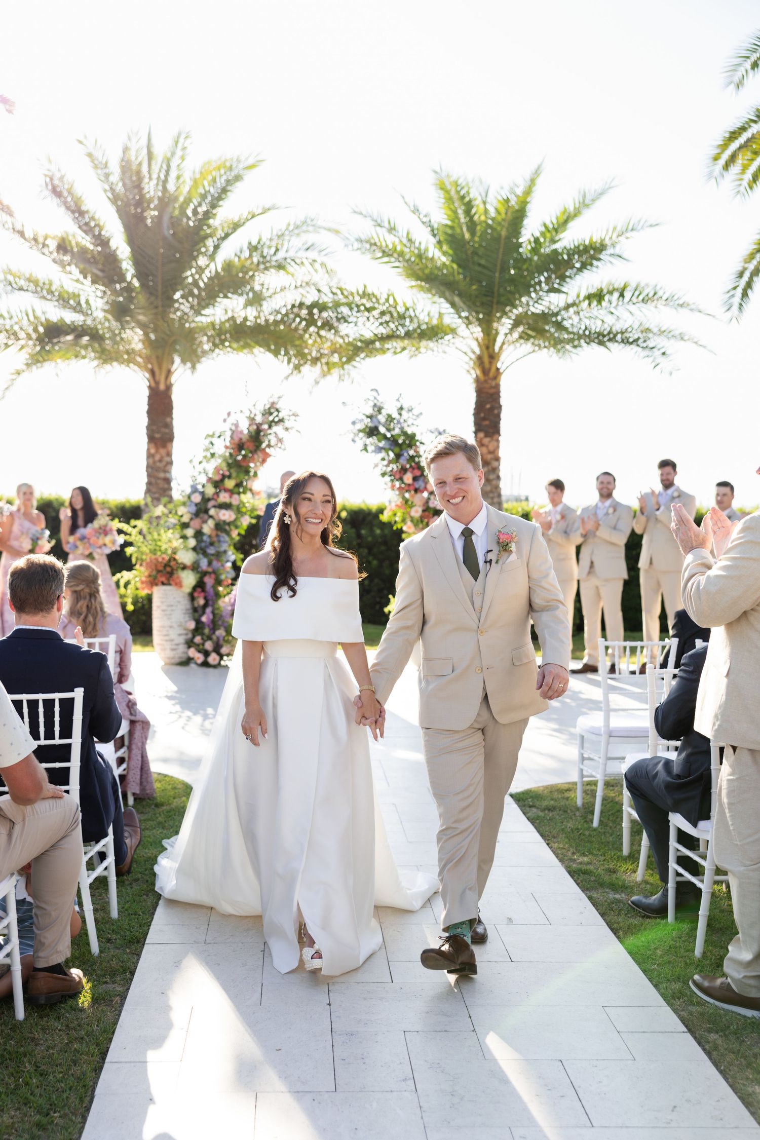 Bride and groom walk down the aisle hand-in-hand at an outdoor wedding ceremony with palm trees.