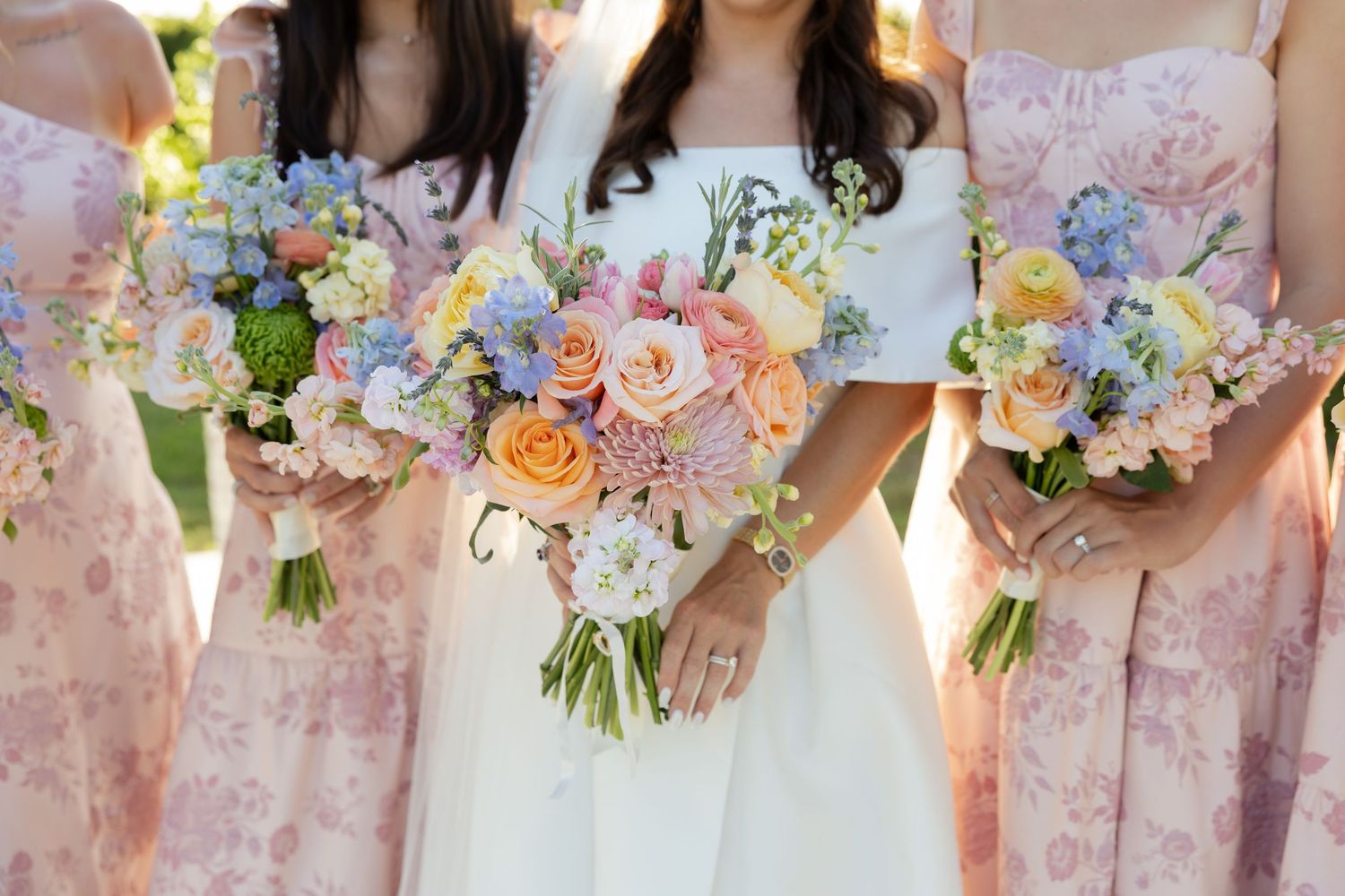 Bride and bridesmaids holding pastel pink, blue, and white floral bouquets with roses at a wedding ceremony.