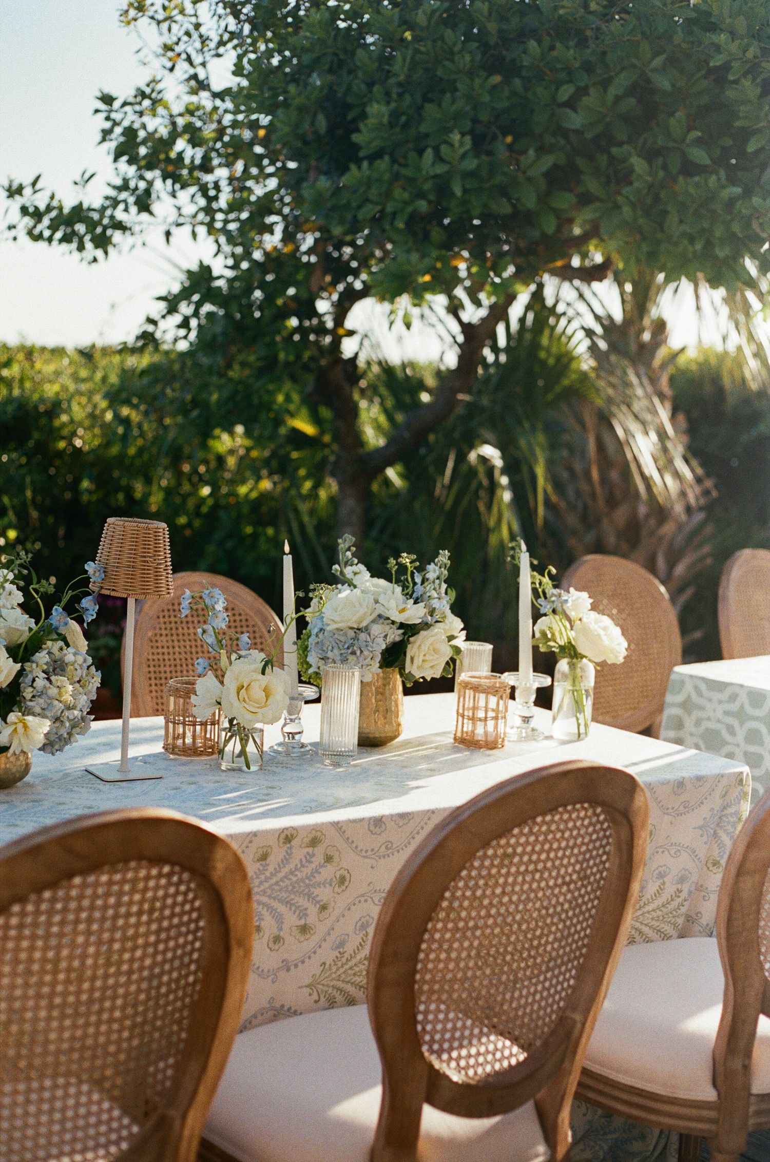 Elegant outdoor wedding table with floral centerpieces, candles, wicker chairs, surrounded by lush greenery documented on film.