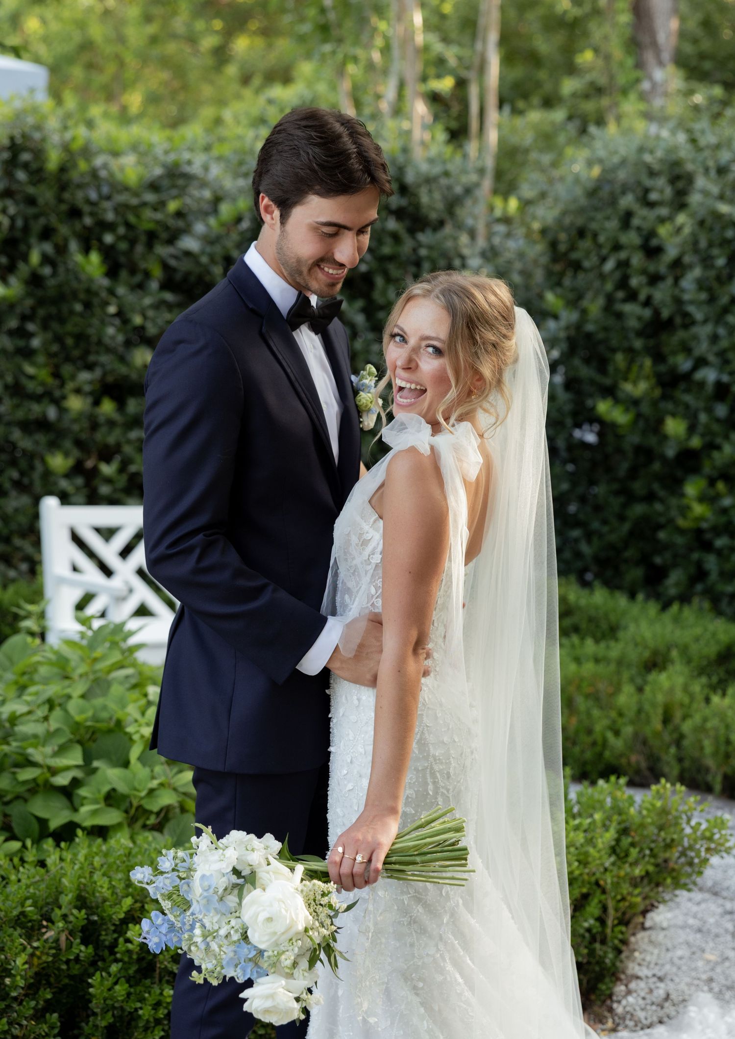 Bride and groom posing in a lush garden, she holds a blue and white bouquet wearing a lace gown with veil.