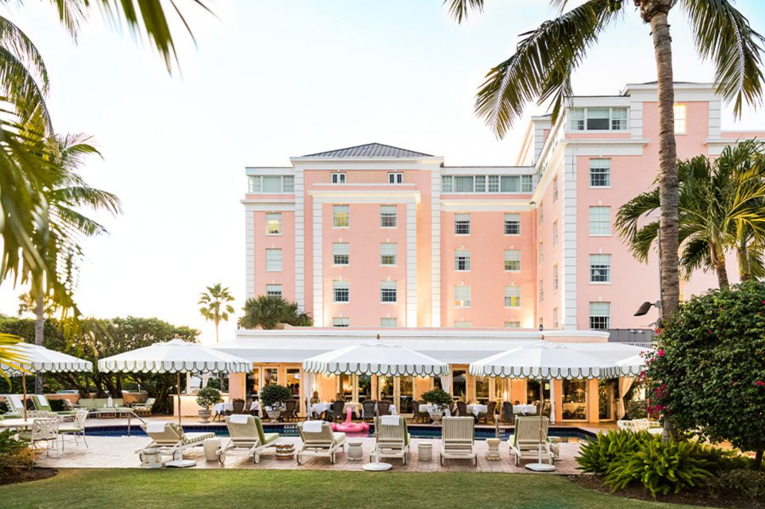 Elegant pink hotel with palm trees, outdoor dining terrace, and white lounge chairs in a tropical setting.