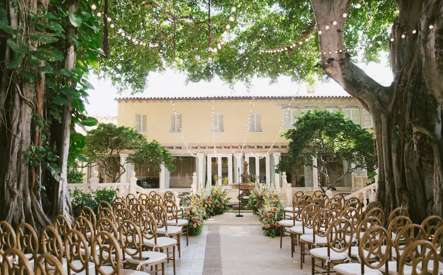 Elegant outdoor wedding ceremony setup with gold chairs, floral aisle, lush trees, and a yellow villa backdrop.