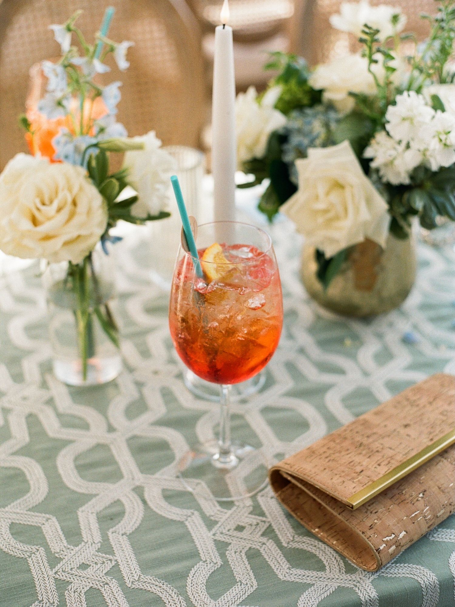 Elegant Aperol Spritz cocktail with blue straw on a sage green tablecloth, surrounded by white roses and candles.
