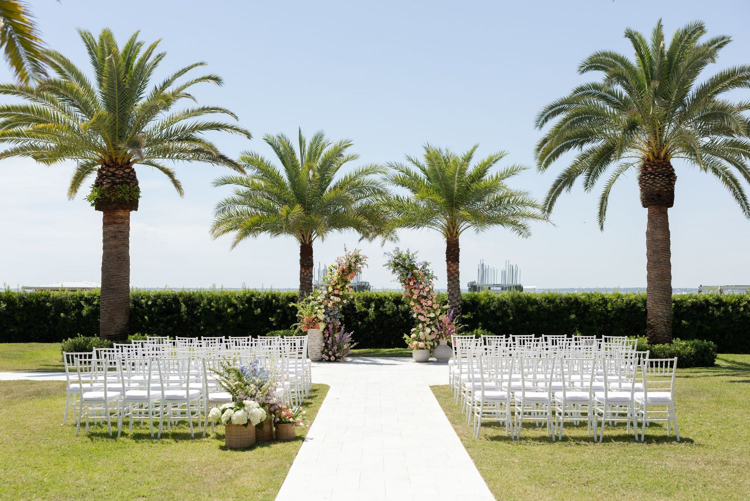 Outdoor wedding ceremony setup with white chairs, floral arch, and palm trees overlooking a scenic waterfront on a sunny day.