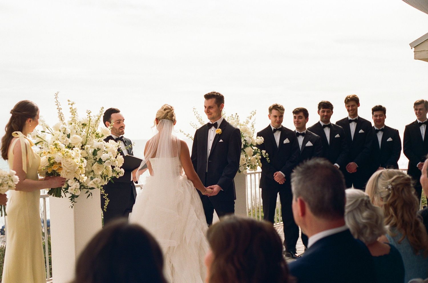 Elegant Florida outdoor wedding ceremony with bride and groom at altar, groomsmen in tuxedos, and white floral arrangements.