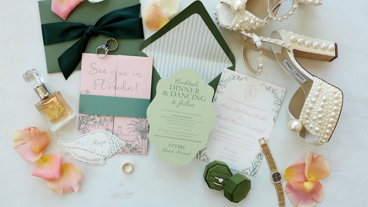 Elegant South Florida wedding flat lay with green invitations, pearl heels, gold perfume, floral accents, and rings on white background.
