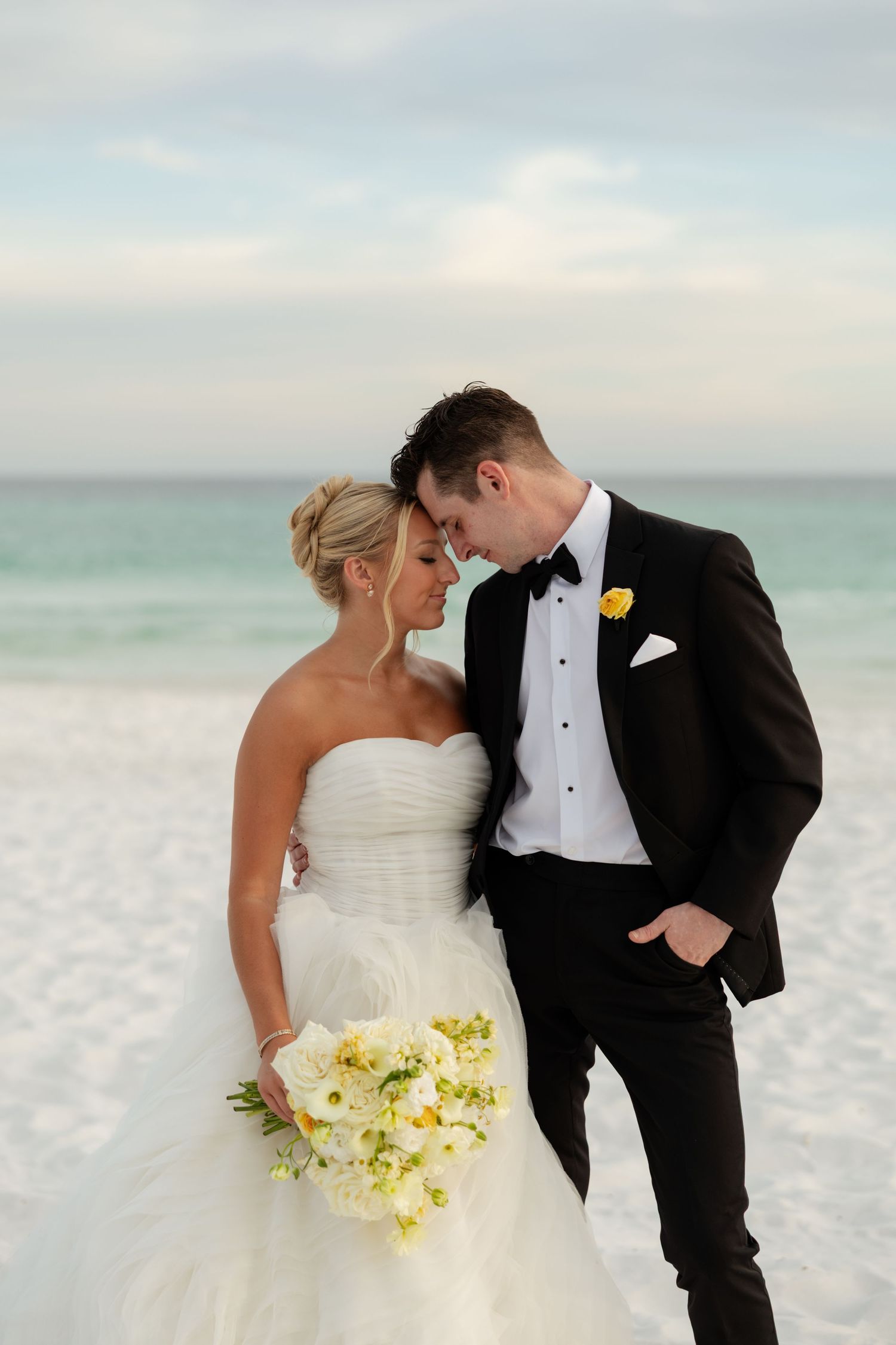 Bride and groom share a romantic moment on a white sand beach, groom in black tuxedo.