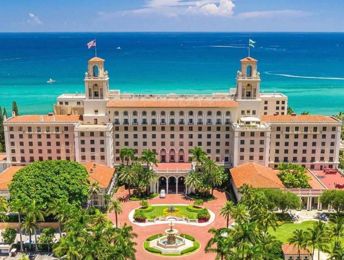 Aerial view of a grand beachfront resort hotel with pink facade, lush gardens, and turquoise ocean backdrop.