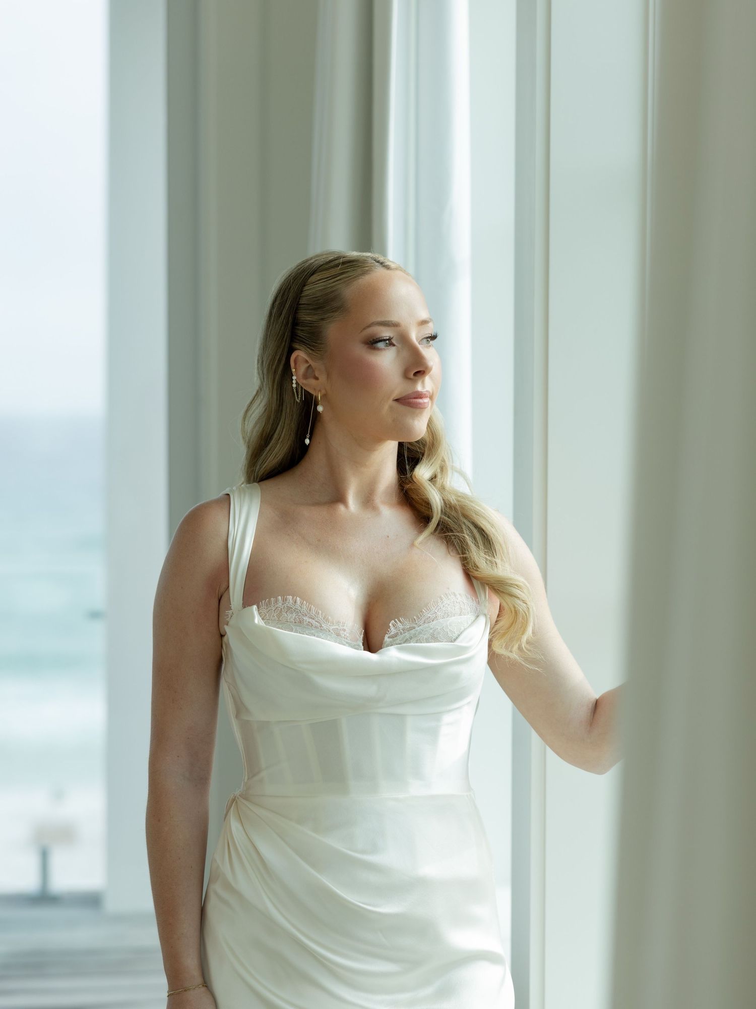 Bride in white satin corset wedding gown standing by large window with ocean view, blonde hair, pearl earrings.