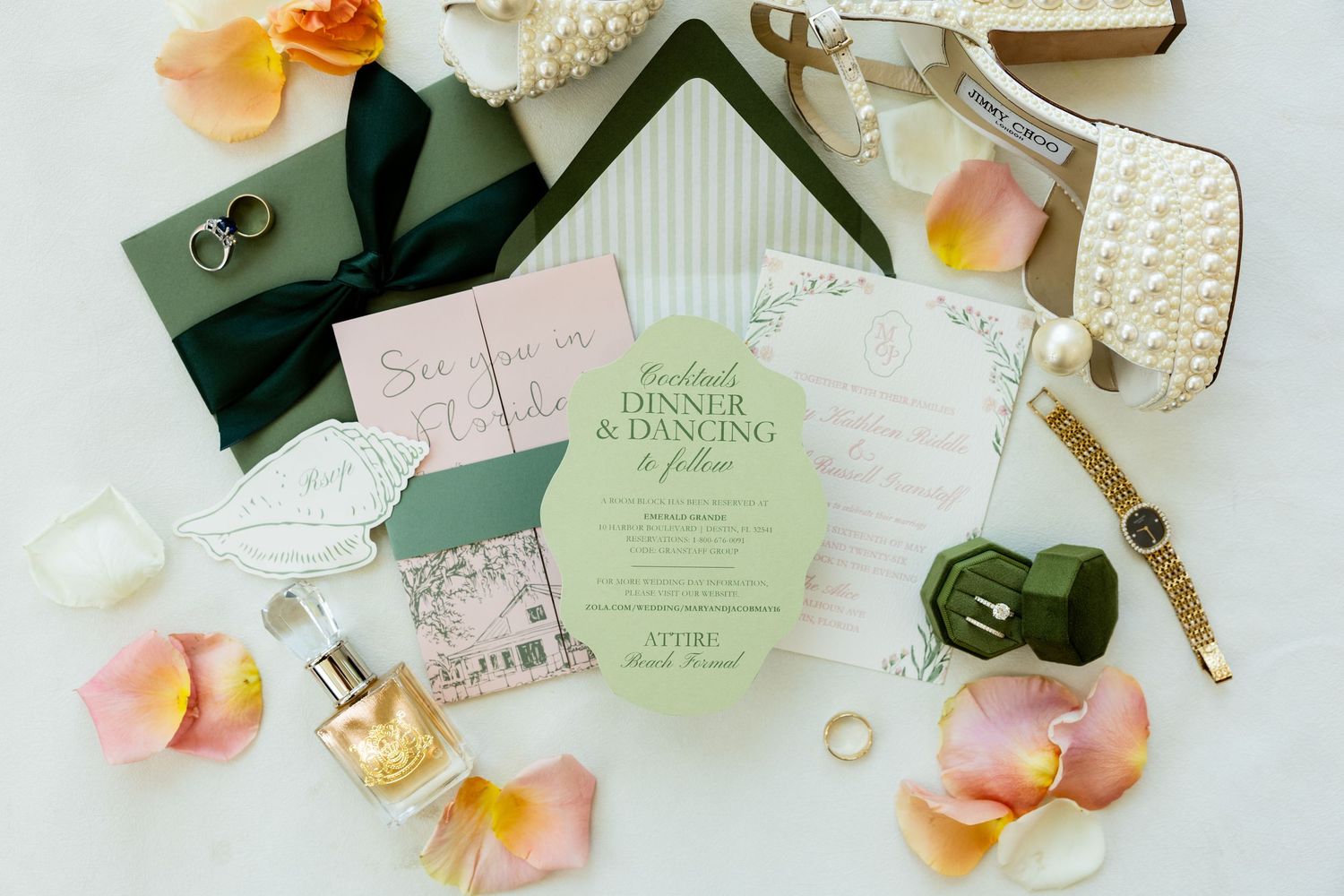 Elegant wedding flat lay with green invitations, pearl heels, floral petals, rings, perfume, and a watch on white background.