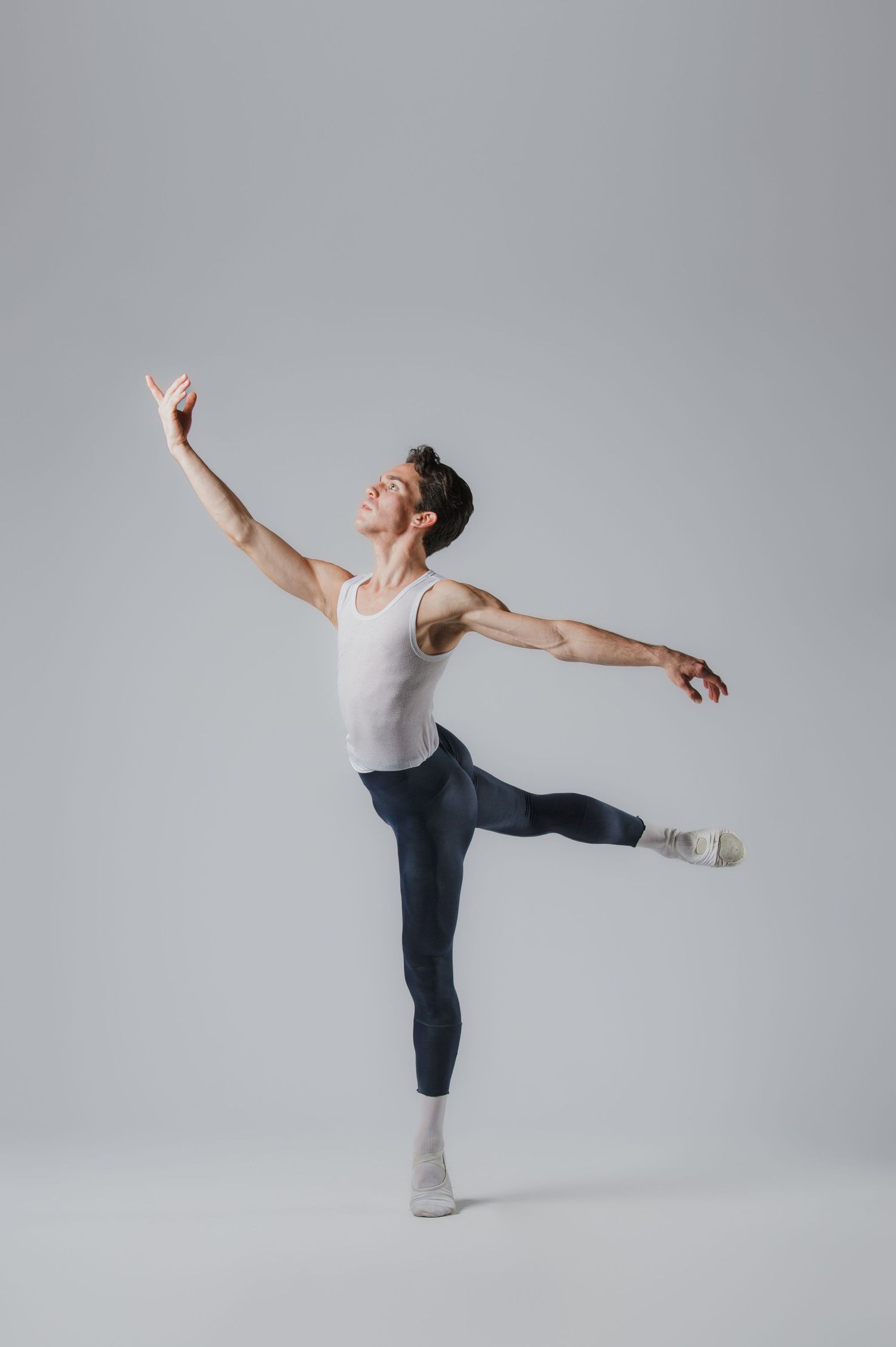 Male ballet dancer in white tank top and blue leggings performing an arabesque pose against gray background.
