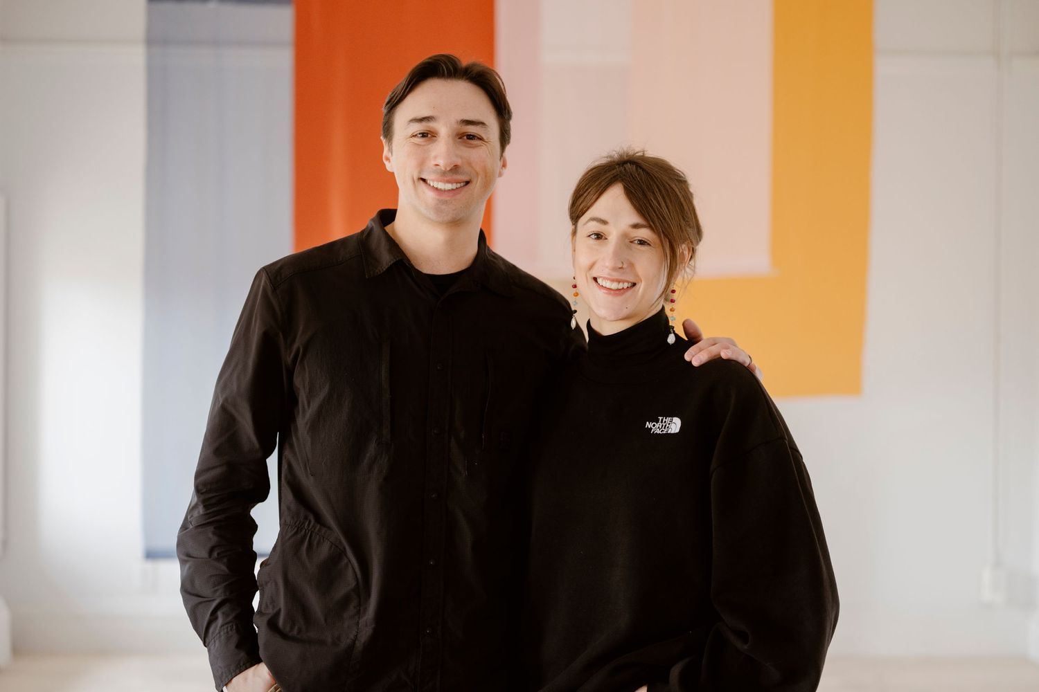 Two people in matching black outfits standing together against an orange and white wall.