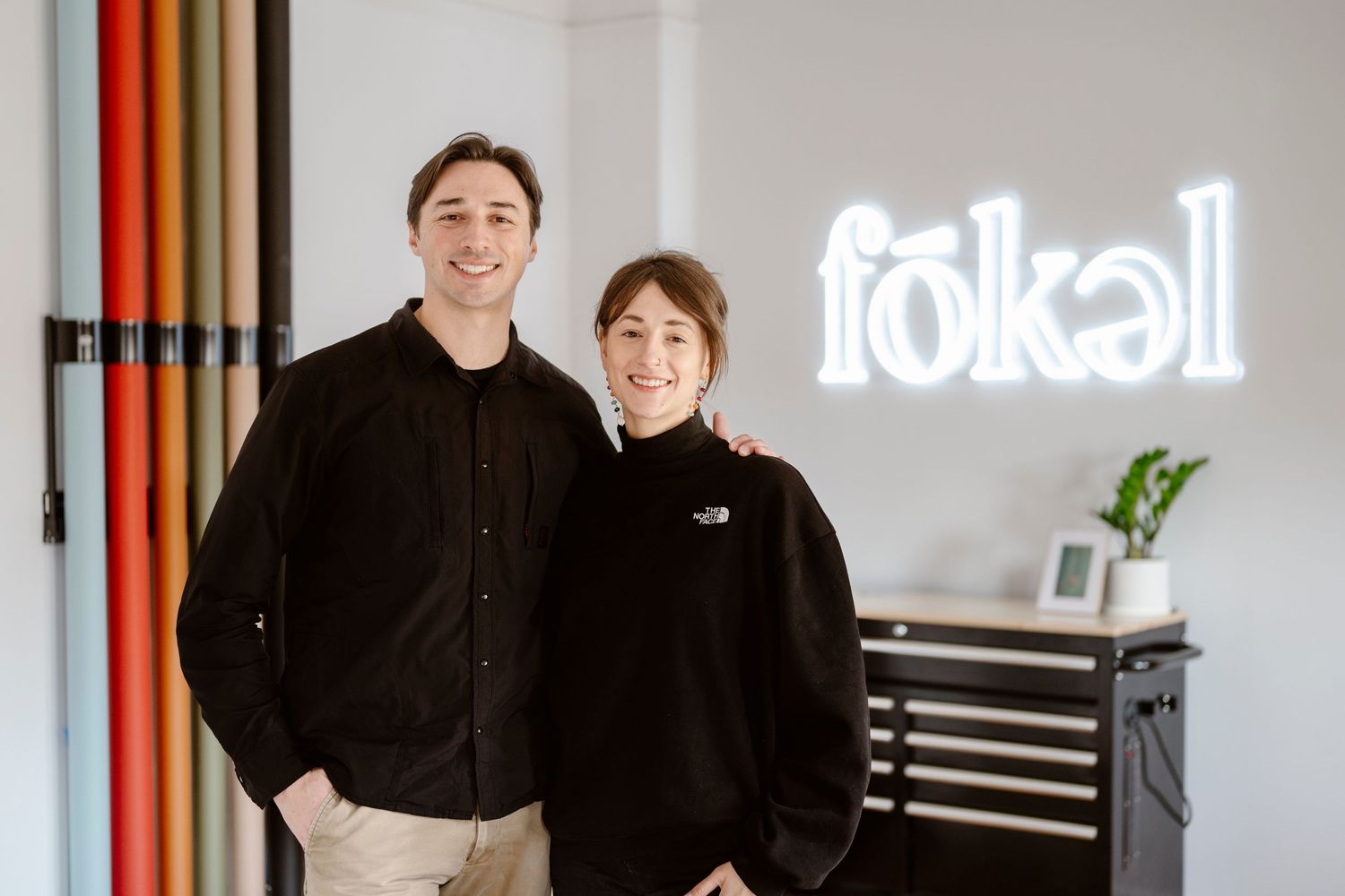 Two people in black attire standing together in a photo studio with neon signage.