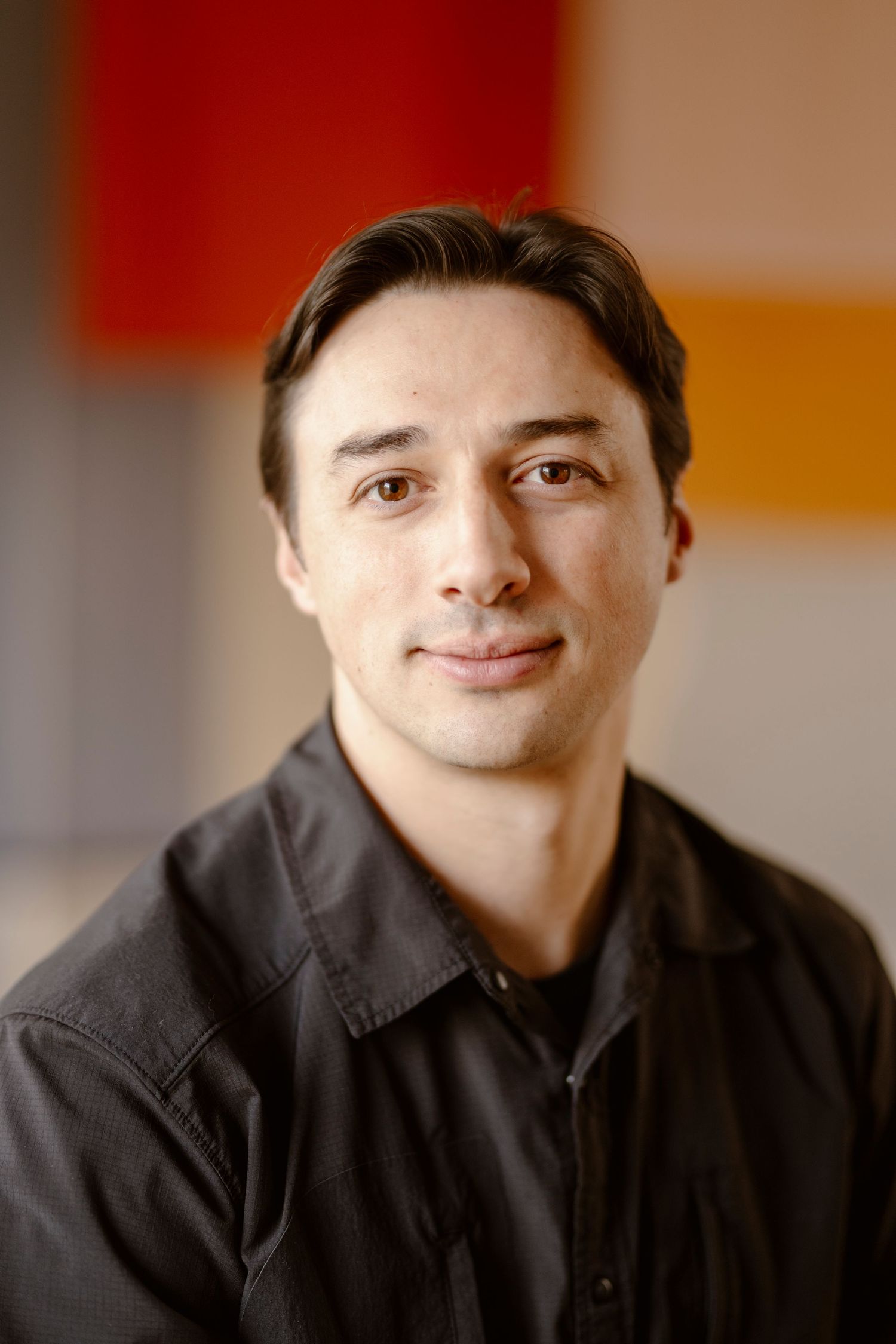 Professional headshot of a person in a dark button-up shirt against a warm-toned background.