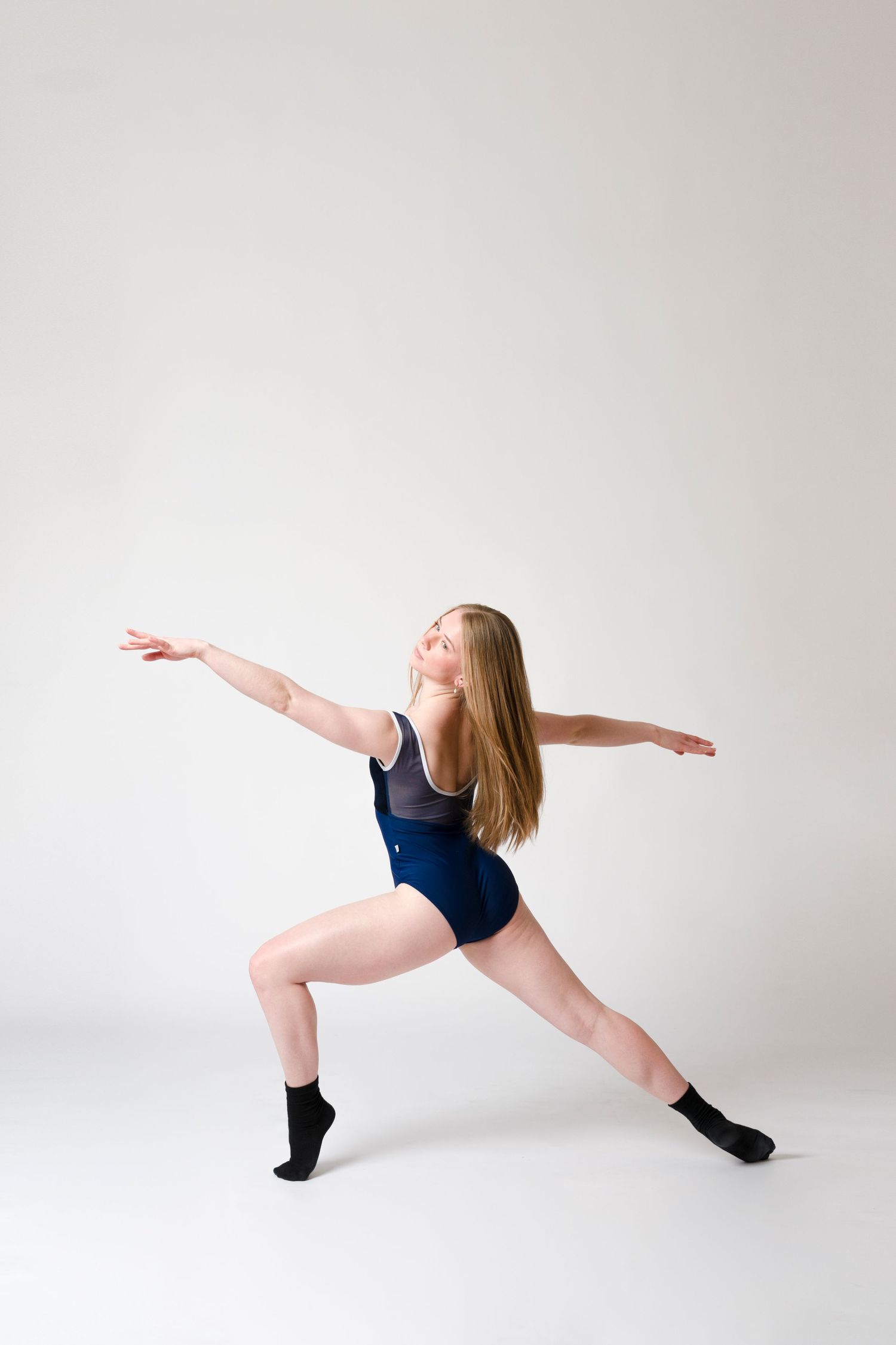 Dancer in navy blue leotard performs graceful lunge pose with outstretched arms against white studio background.