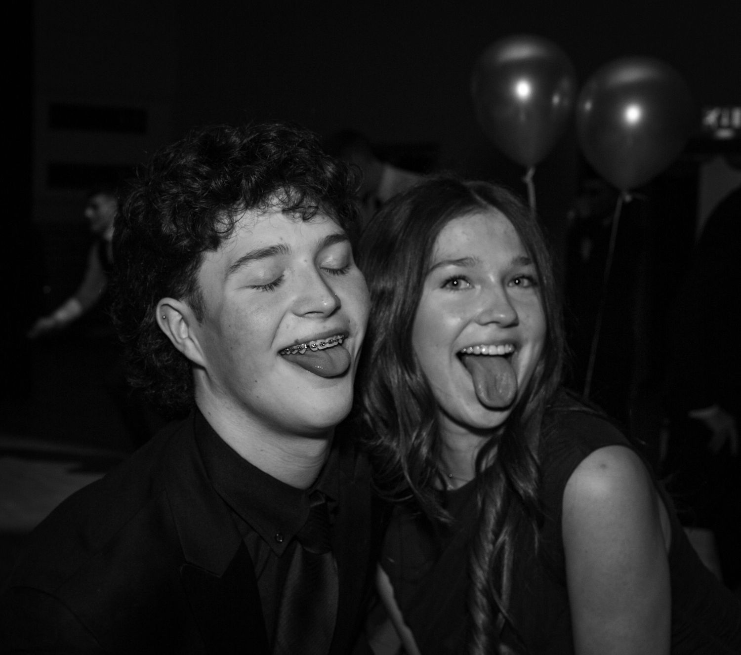 people having fun sticking their tongues out for the camera at a winter ball event in the hall of a school in Edinburgh in black and white.