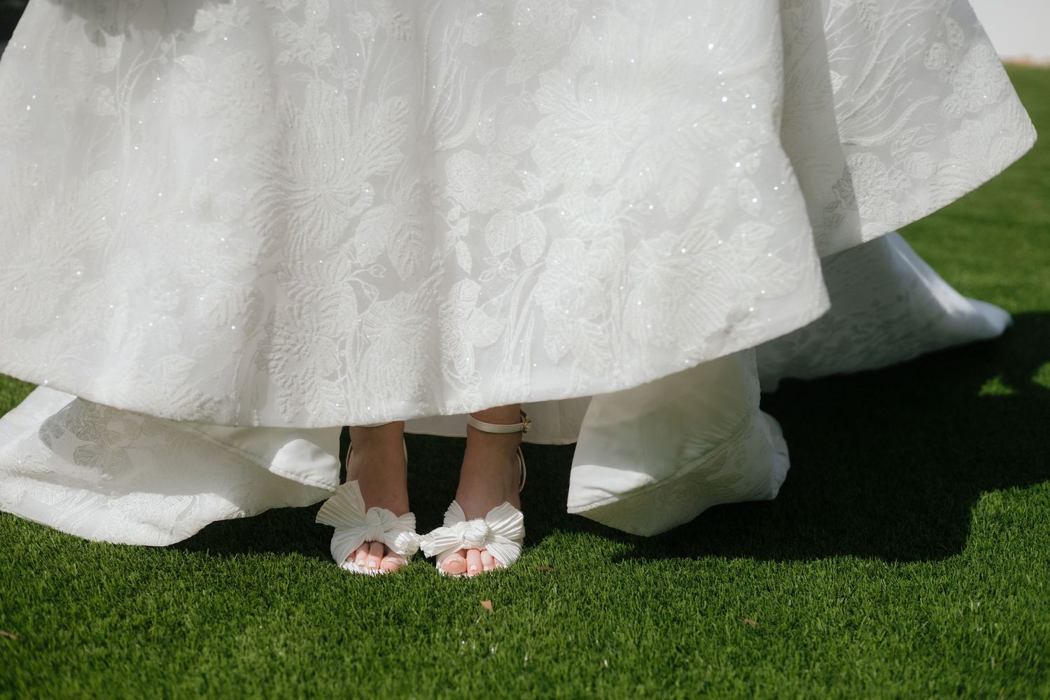 A row of wedding dress hems and white bridal shoes shown against green grass in an outdoor setting.