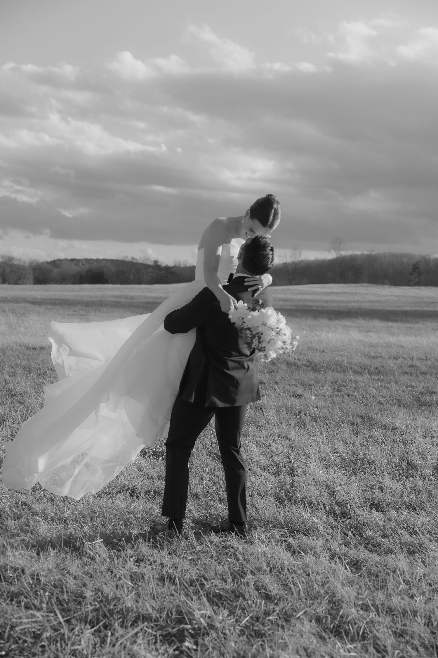 A romantic black and white wedding photo showing a couple embracing in a scenic field under dramatic clouds.