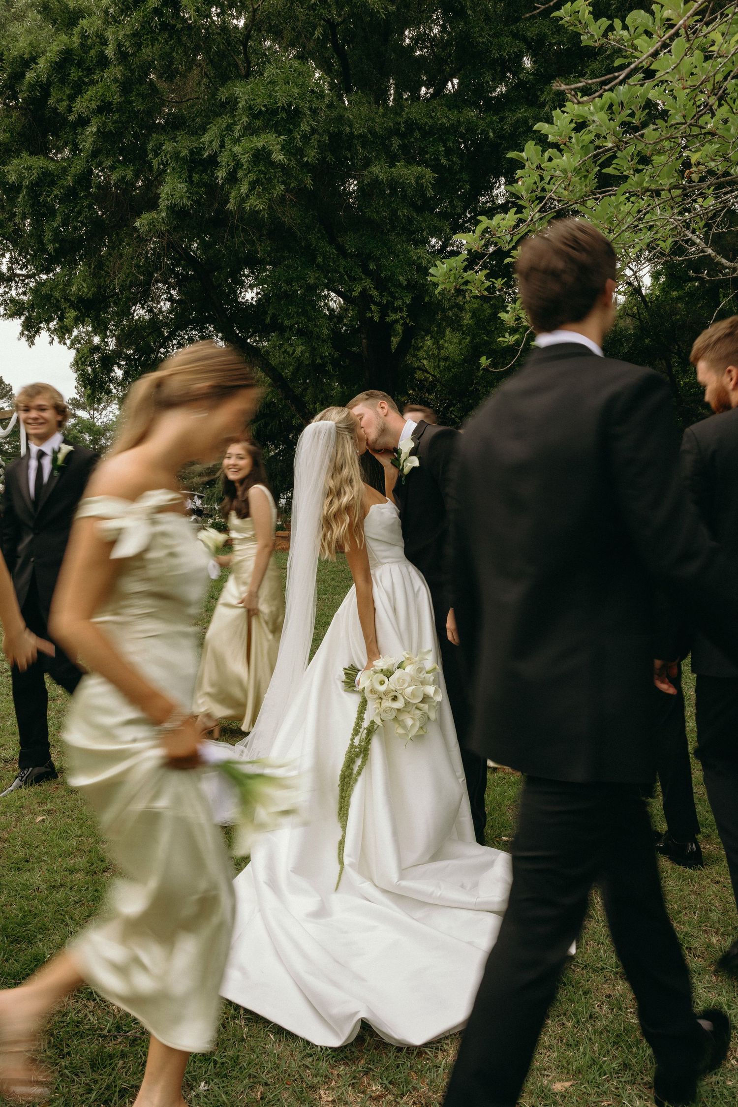Bride and groom share a kiss surrounded by wedding party in elegant attire outdoors near lush green trees.