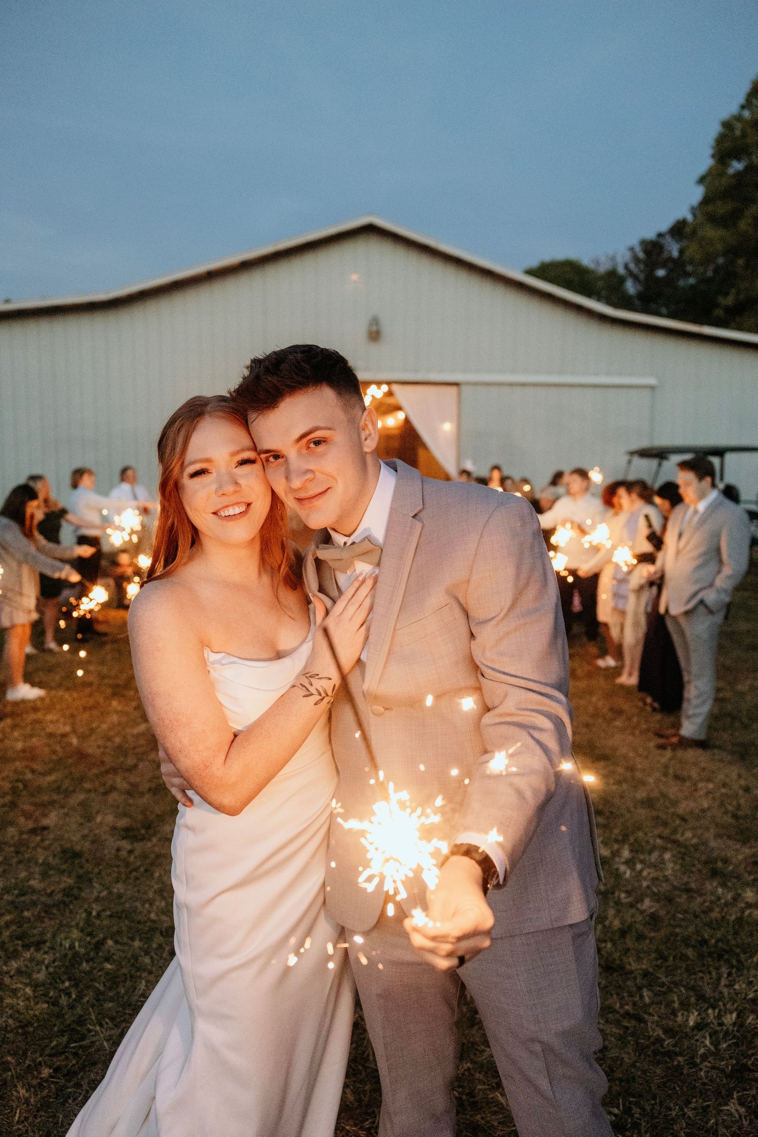 Wedding couple holds sparklers outside barn at twilight with guests in background.