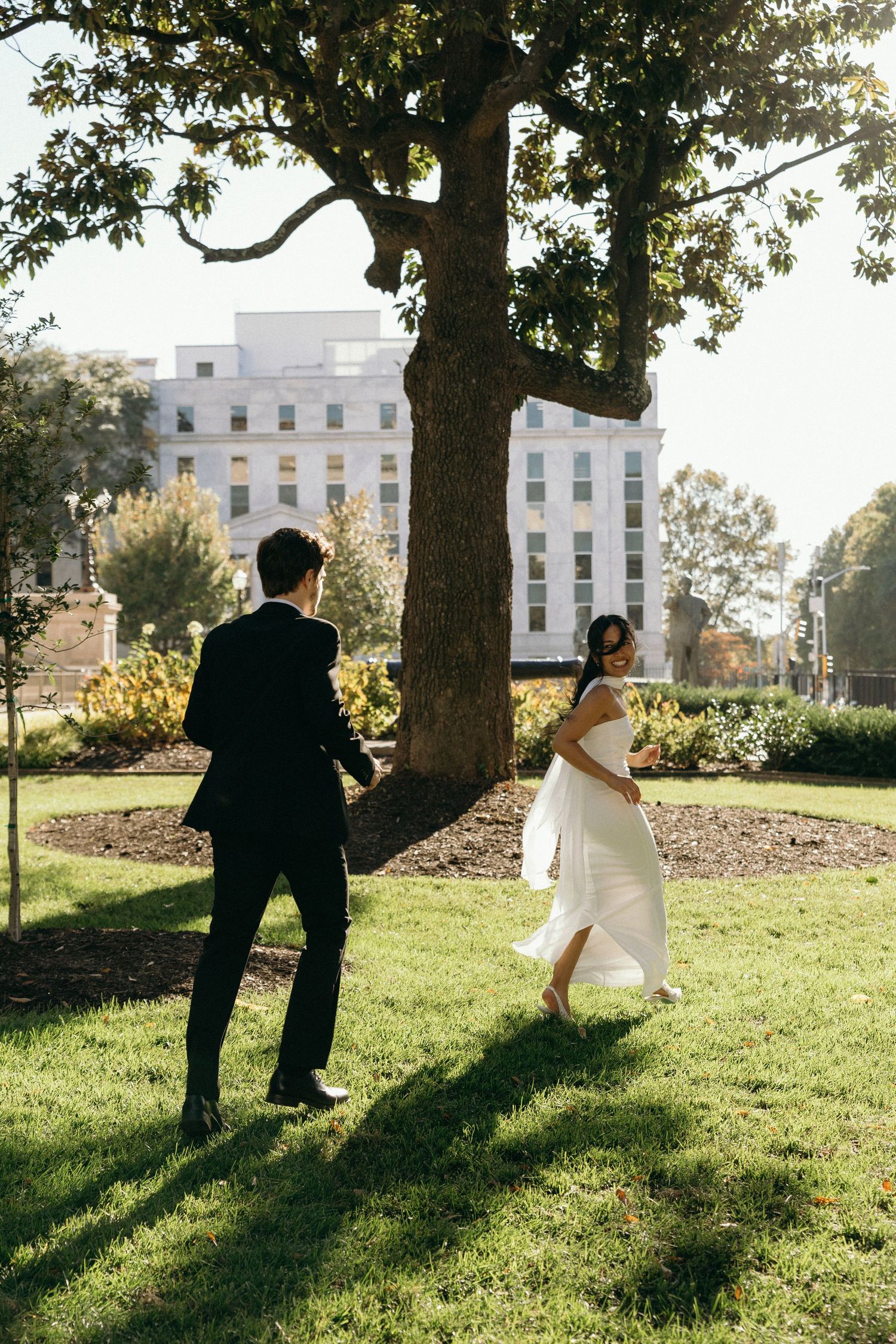 A bride and groom walk through a sunlit garden with a grand white mansion in the background.
