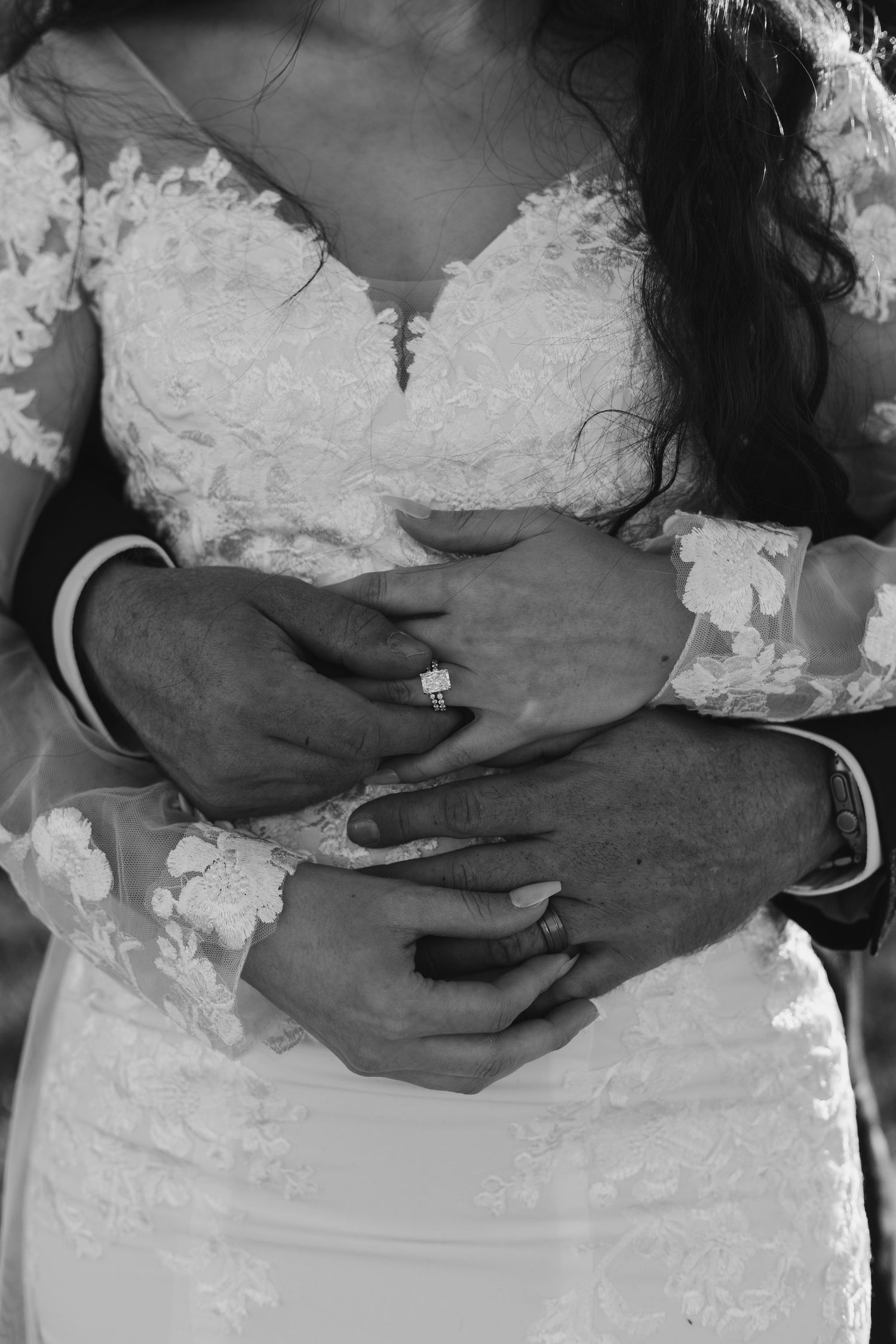Close-up black and white photo of hands with wedding rings against lace wedding dress.