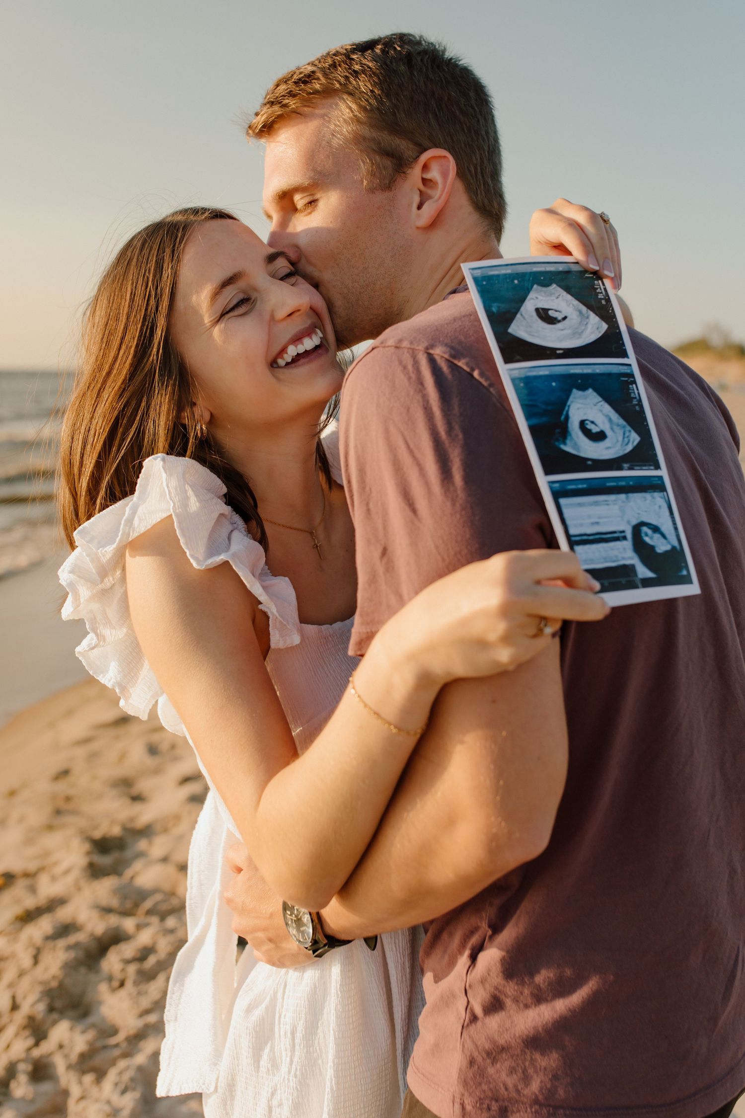 Couple embracing and smiling as they announce their pregnancy on the beach with an ultrasound photo.