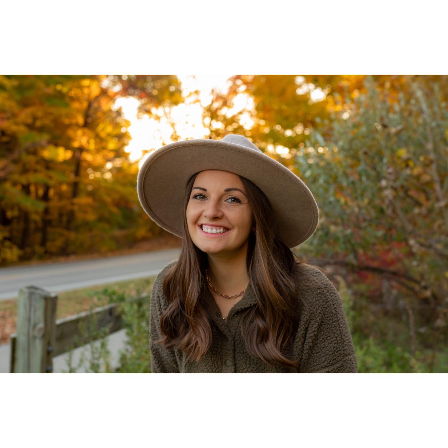 Sarah Prins wearing a hat and sherpa sweatshirt smiling in front of golden fall colors of leaves on trees.