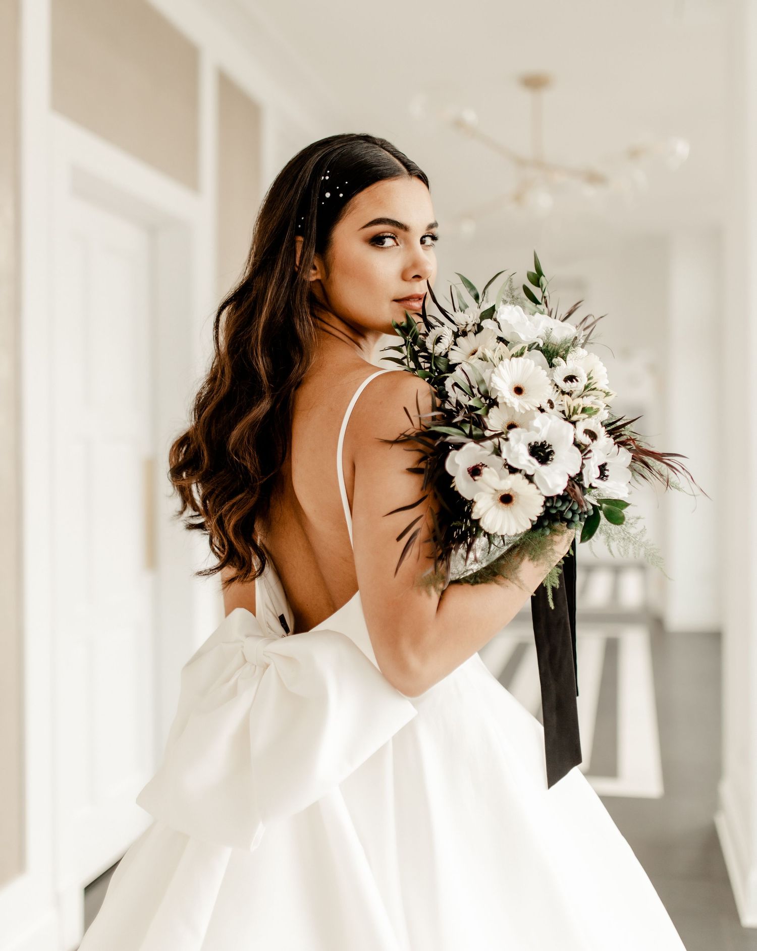 A bride in a white backless gown holds a stunning bouquet of white flowers with trailing black ribbons.