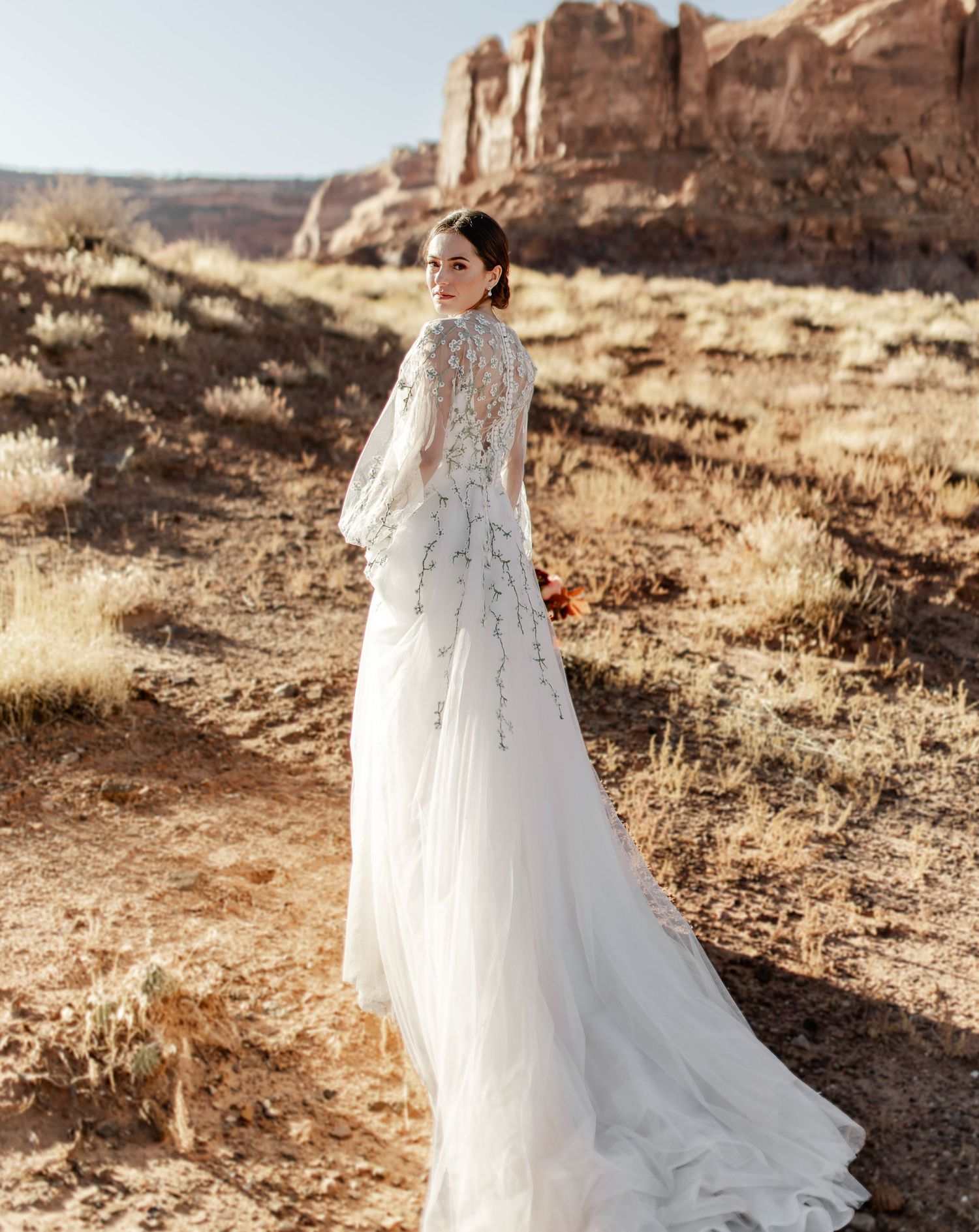 Bride in flowing embellished gown poses against dramatic red rock desert landscape.