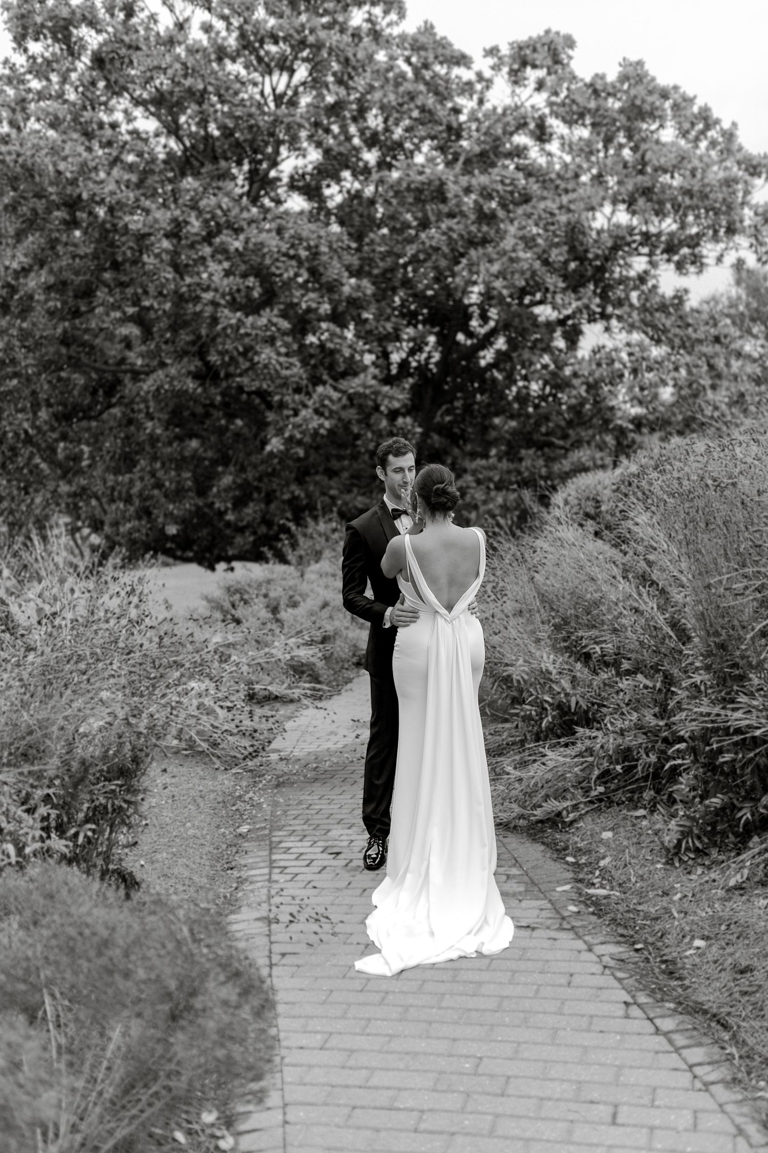 A black and white photo of a couple in formal attire standing on a brick path surrounded by garden foliage.