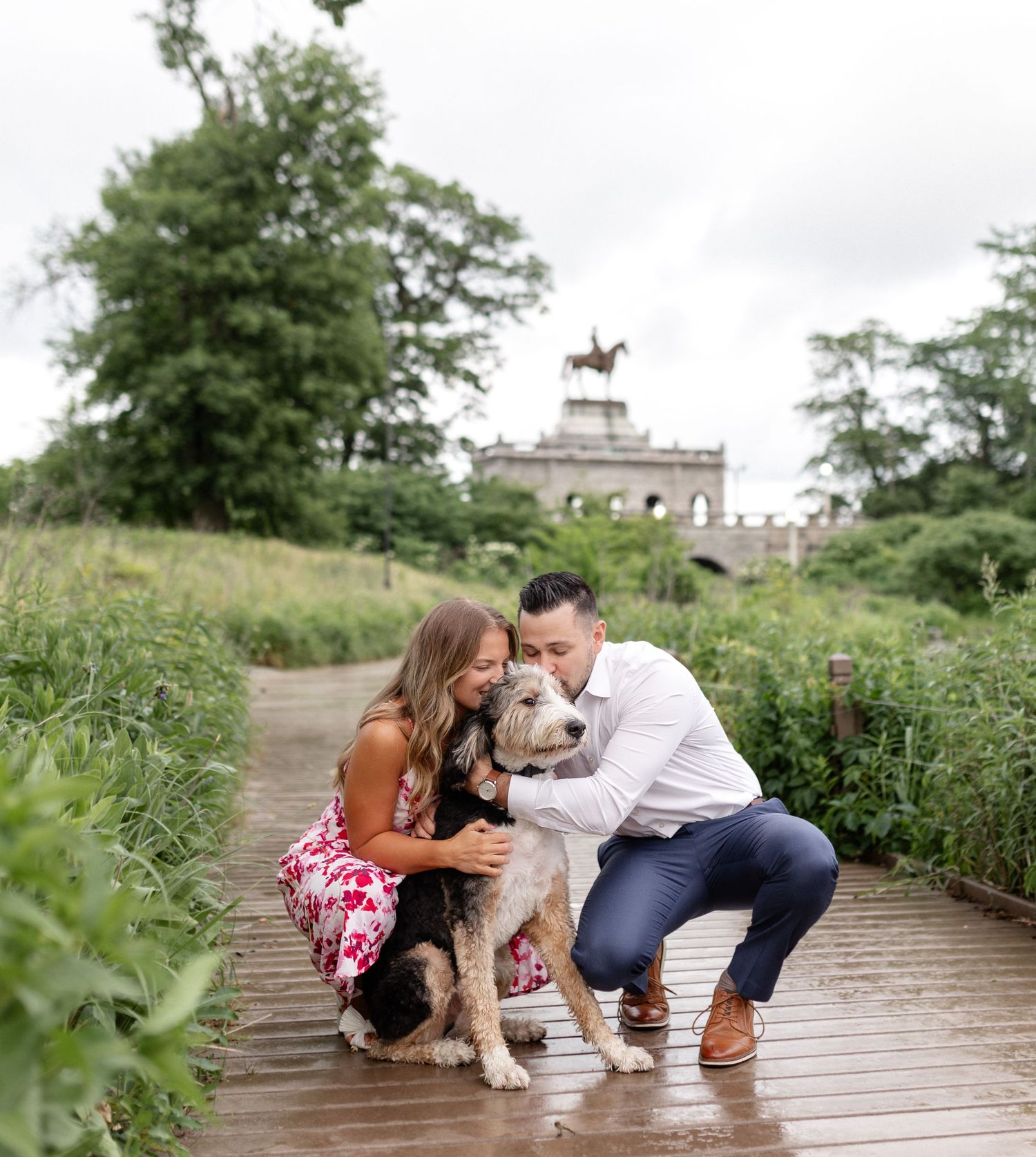 Couple crouching on a wooden path with their fluffy dog, surrounded by lush greenery and a stone monument in the background.