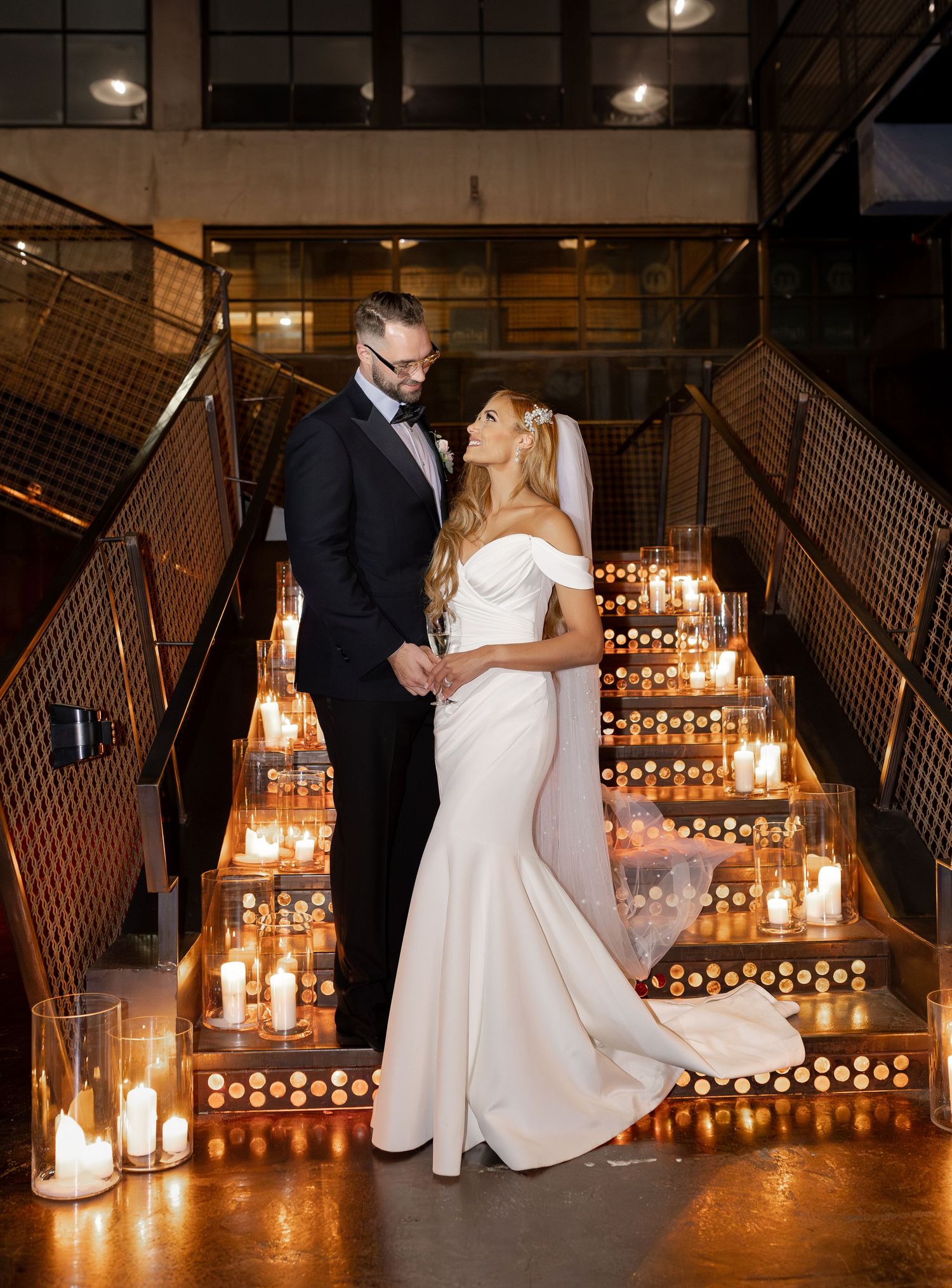 A couple poses on candlelit stairs in formal attire at an industrial-style wedding venue.