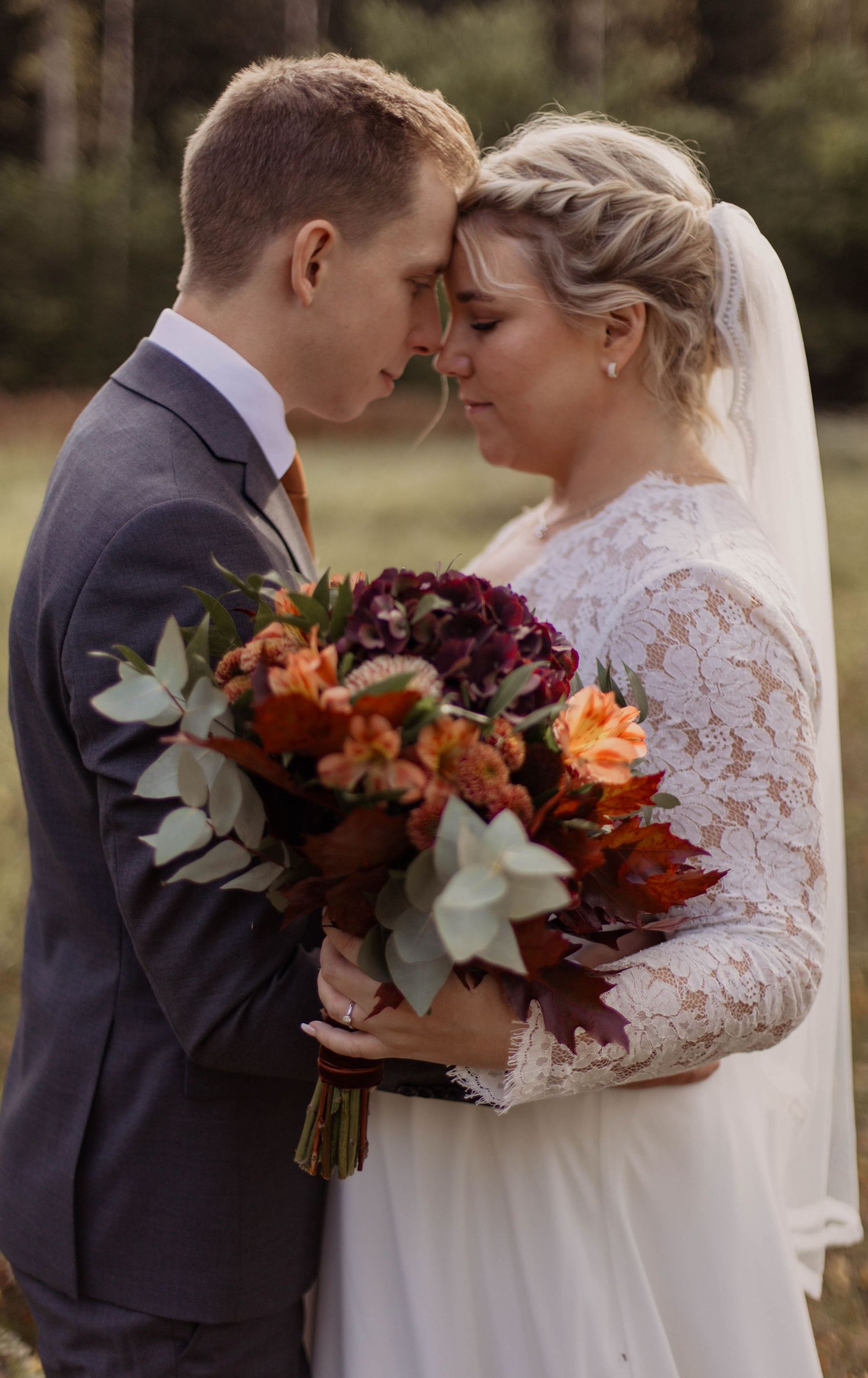 Bride and groom touching foreheads tenderly, holding autumn bouquet with orange and burgundy flowers, outdoors in fall setting.