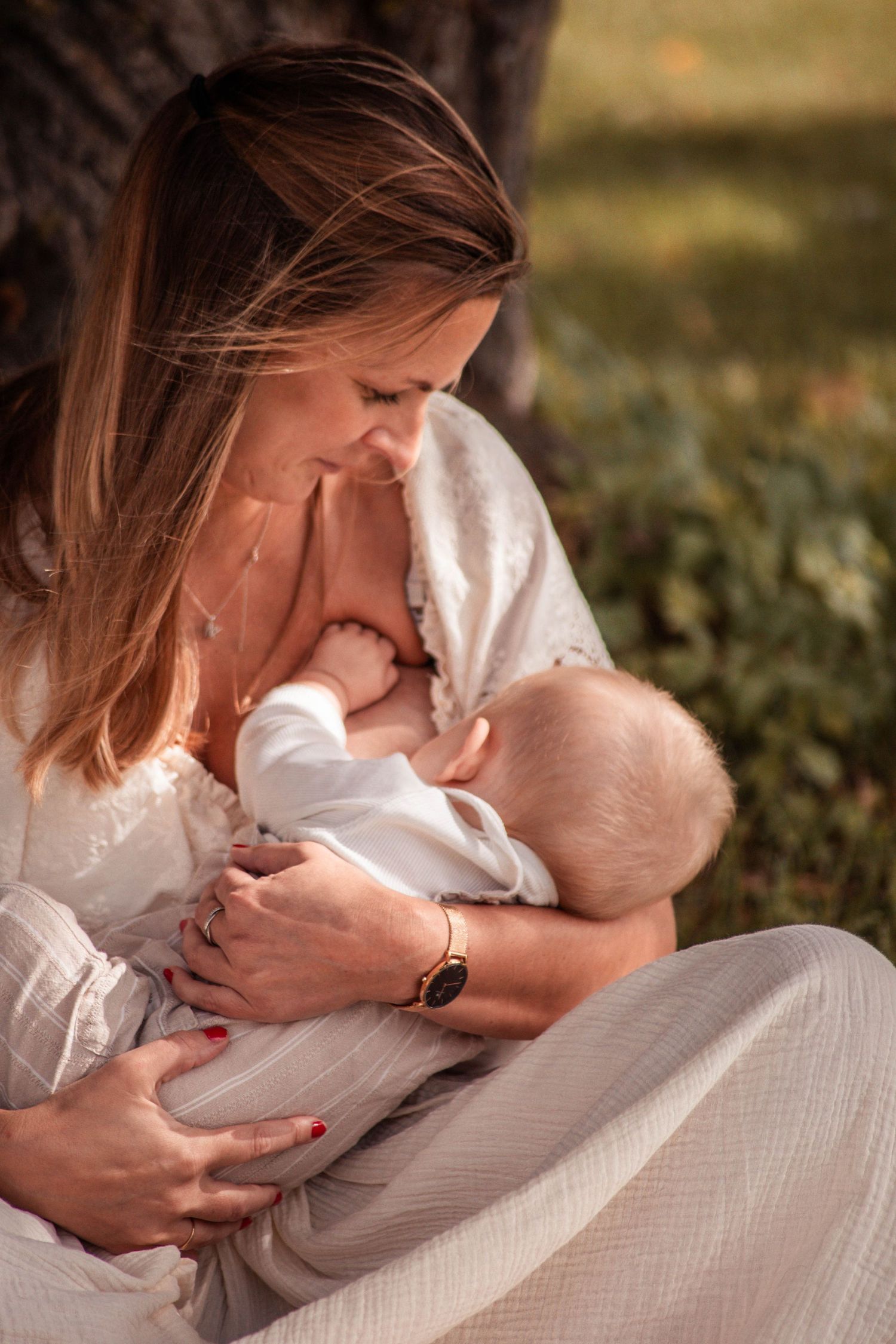 A mother tenderly breastfeeds her infant outdoors, both dressed in white, bathed in warm golden sunlight.
