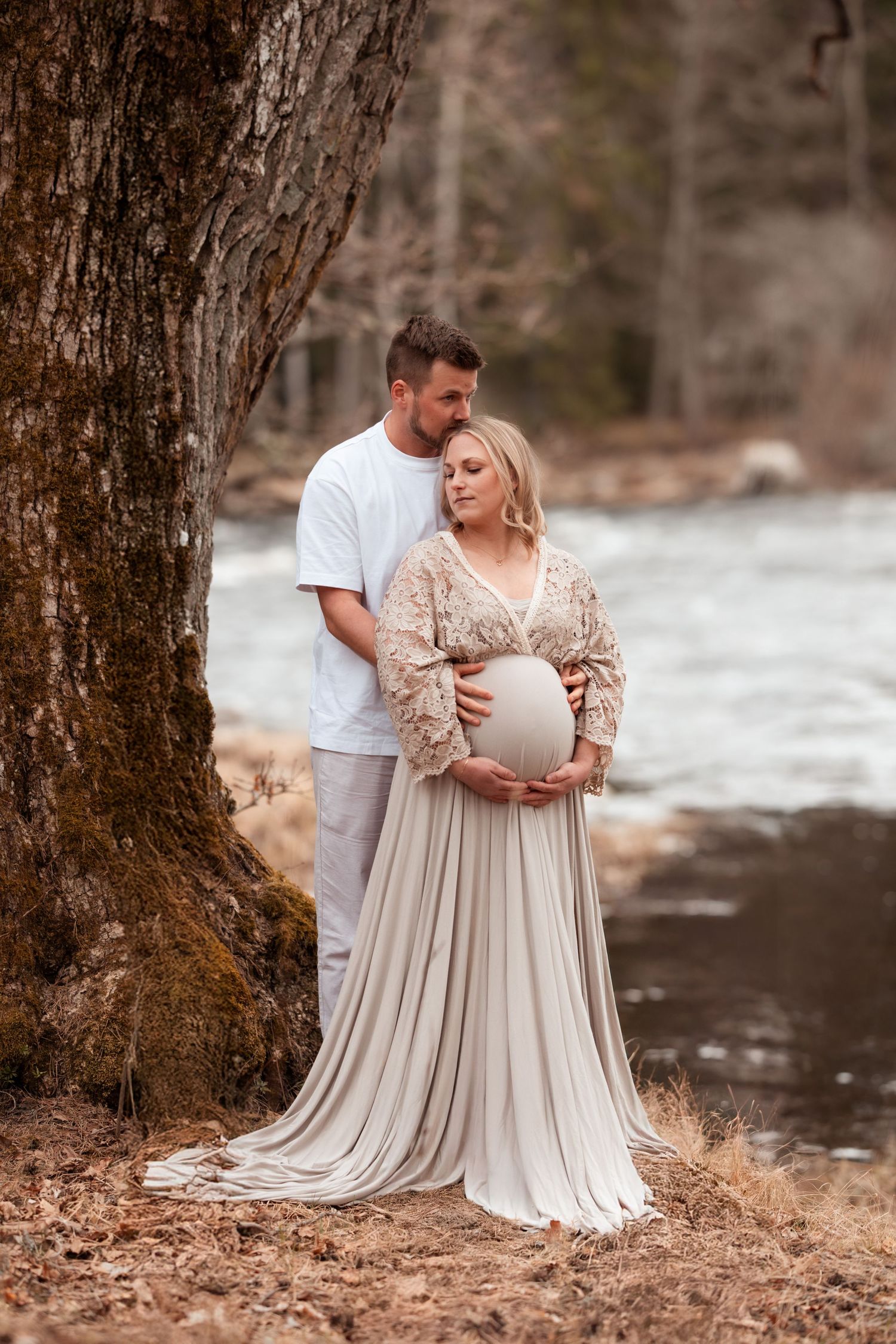 Couple poses by river tree, pregnant woman in flowing beige gown cradling bump, man in white shirt embracing her.