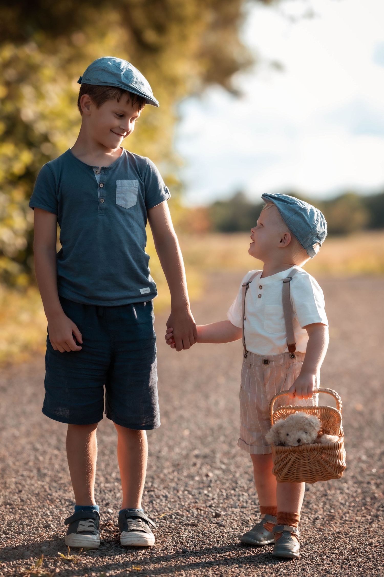 Two young brothers in matching caps hold hands on a dirt road, the younger carrying a wicker basket with a toy.