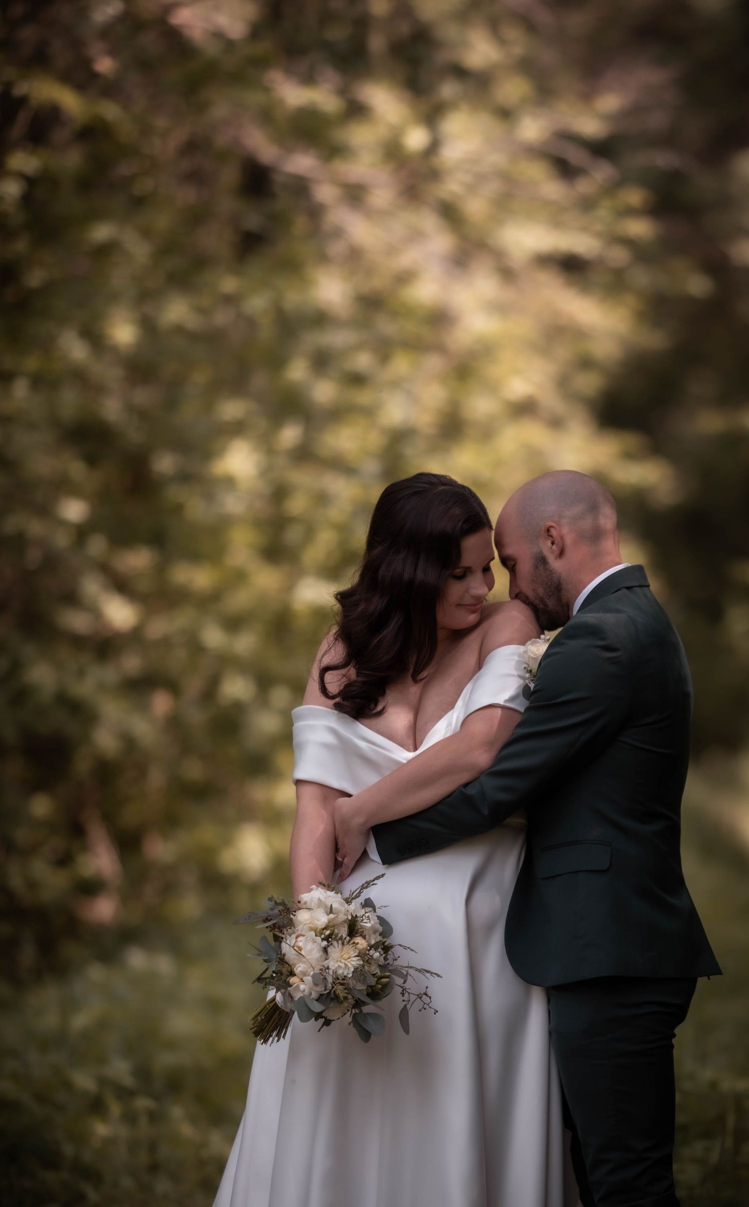 Bride and groom embrace tenderly in a lush forest setting, she holds a white bouquet in an off-shoulder gown.