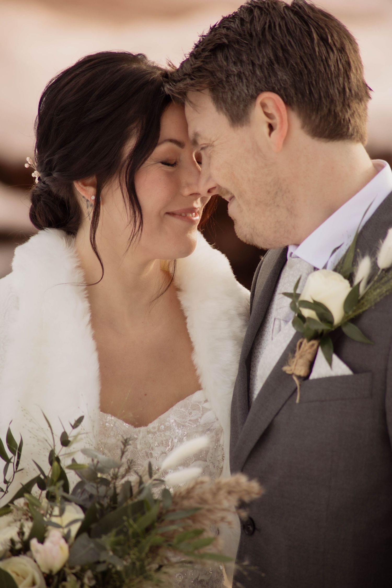 A bride and groom share a romantic moment, touching foreheads, as she holds a lush bouquet of white flowers and greenery.