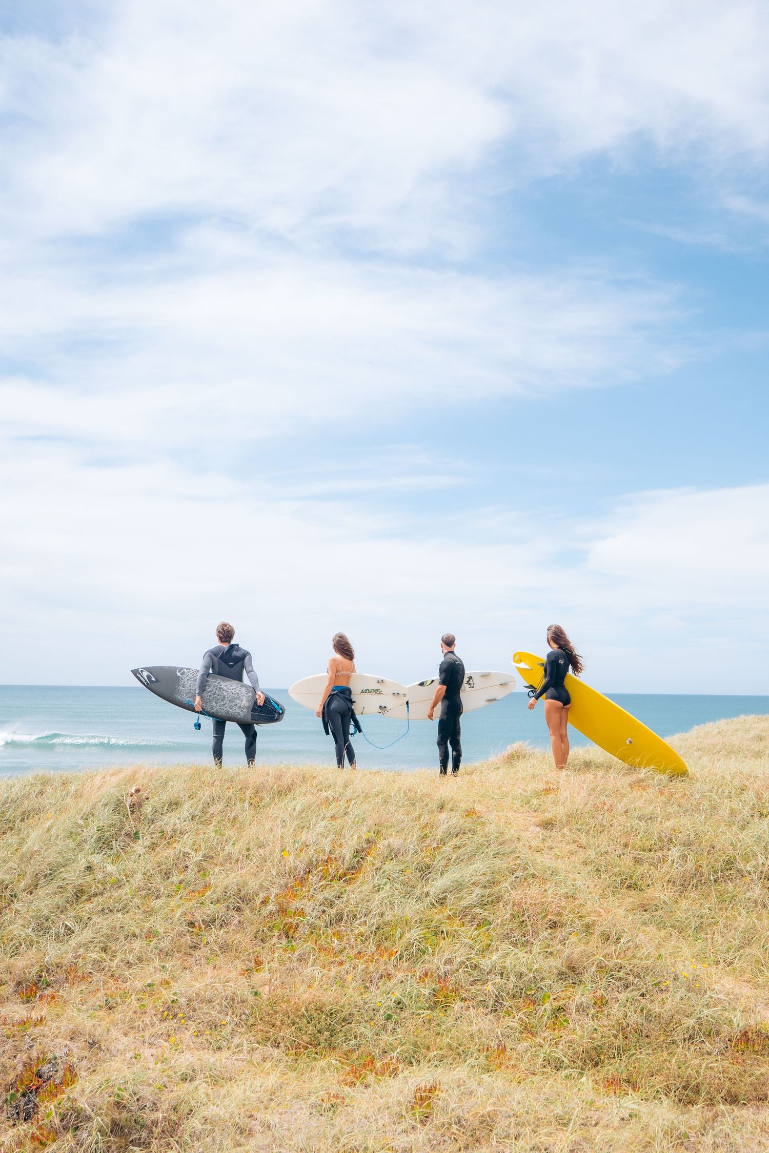 Surfers carrying colorful surfboards walk along a grassy coastal hill overlooking the ocean on a sunny day.