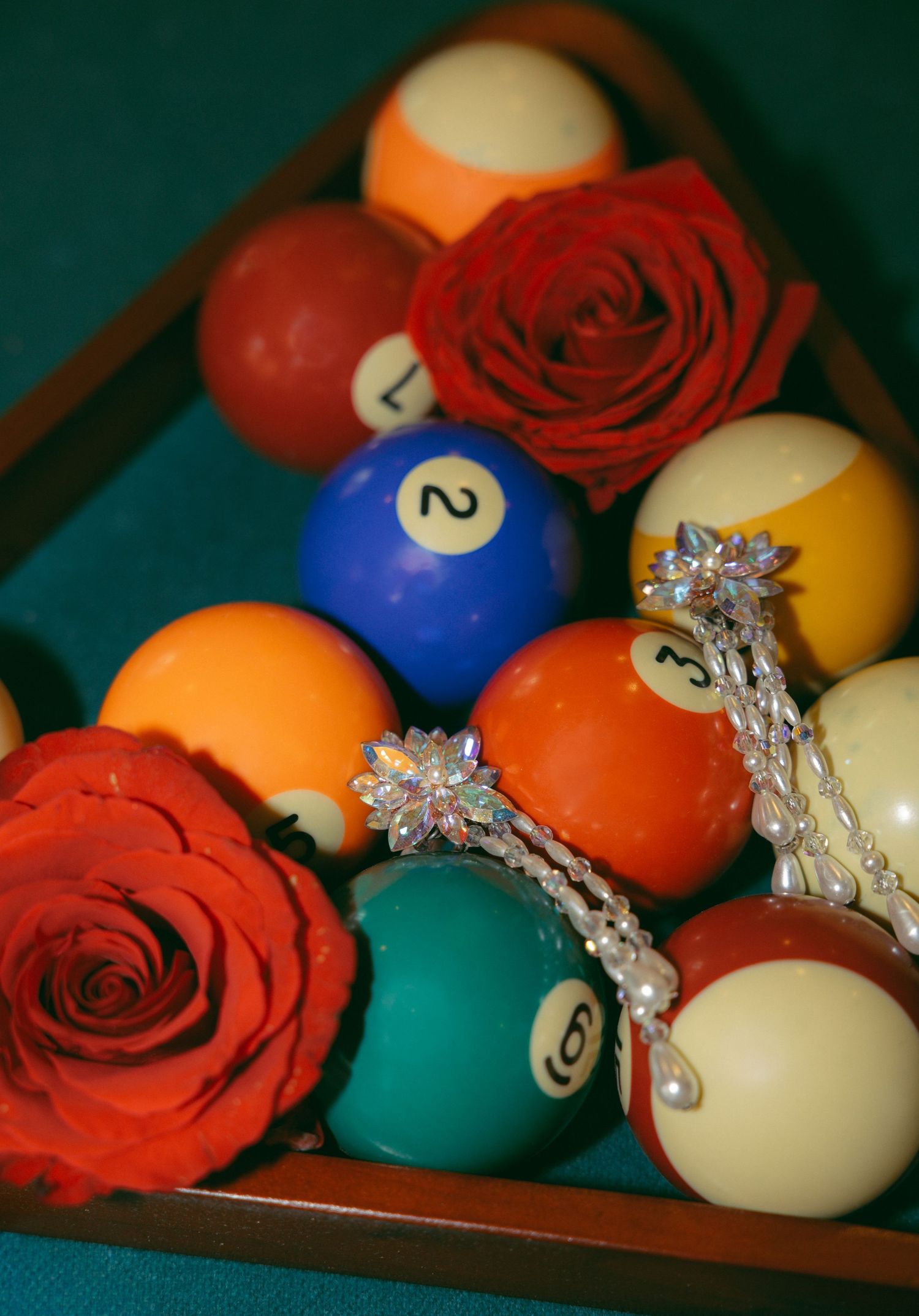 Billiard balls arranged in a rack on a green felt table, decorated with red roses and sparkling crystal jewelry.