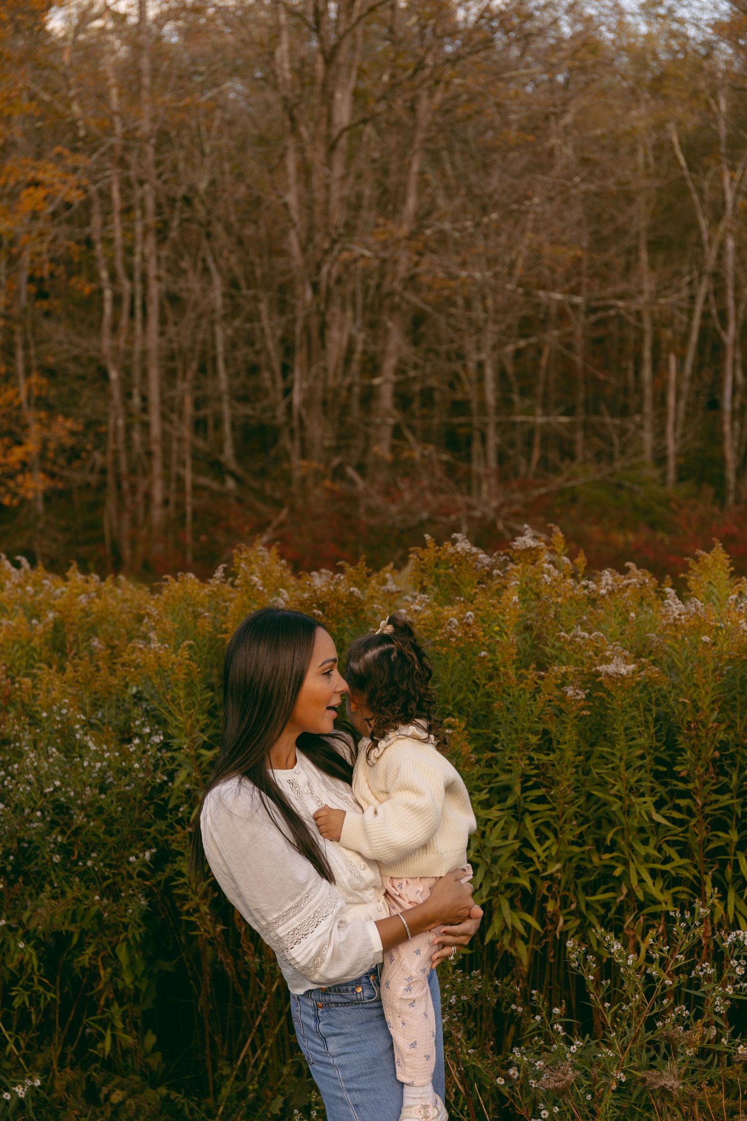 A mother holds her toddler in an autumn field with colorful foliage and trees in the background.