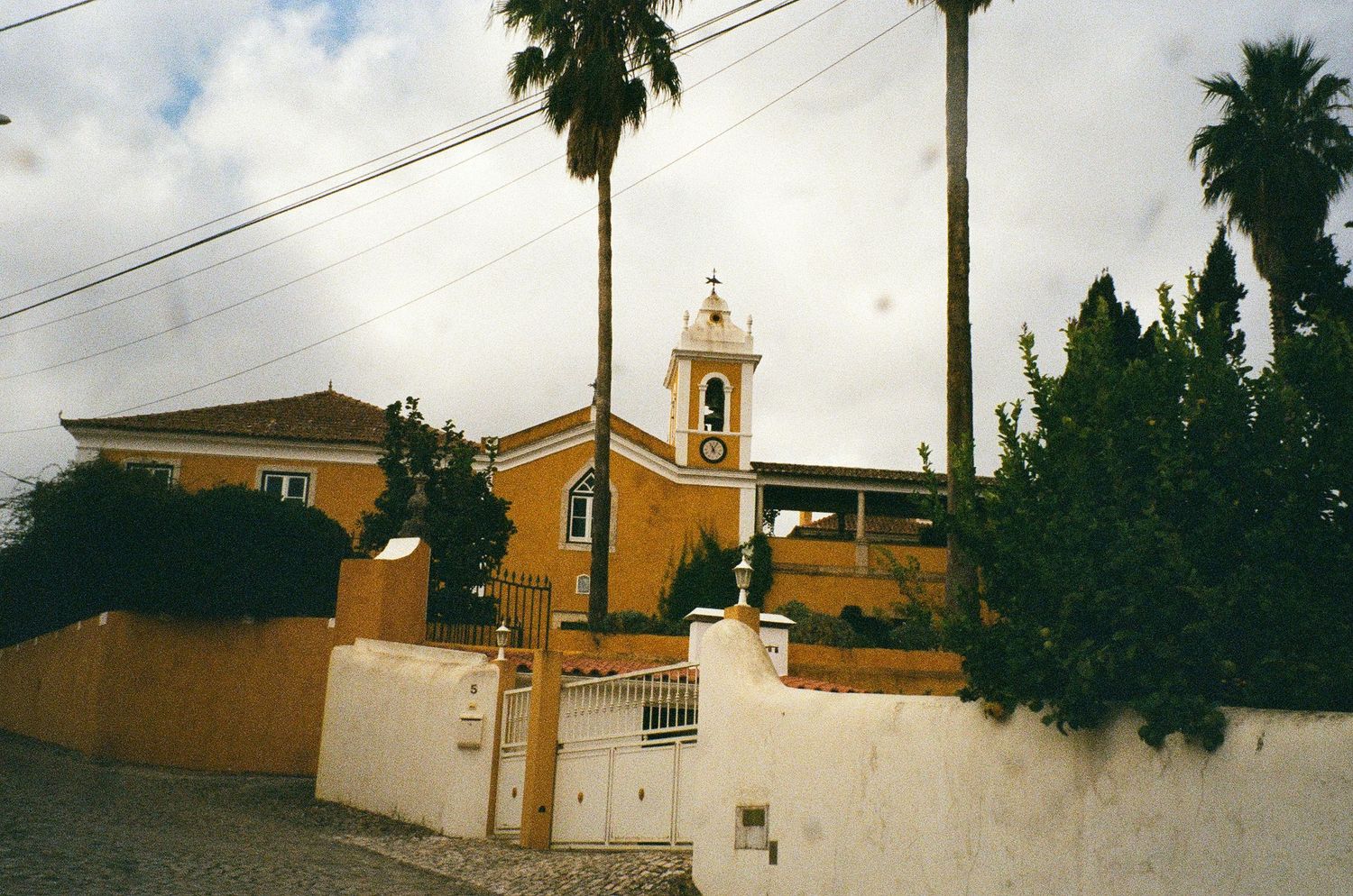 Yellow church with bell tower and palm trees under cloudy sky in a sunny Mediterranean village setting.
