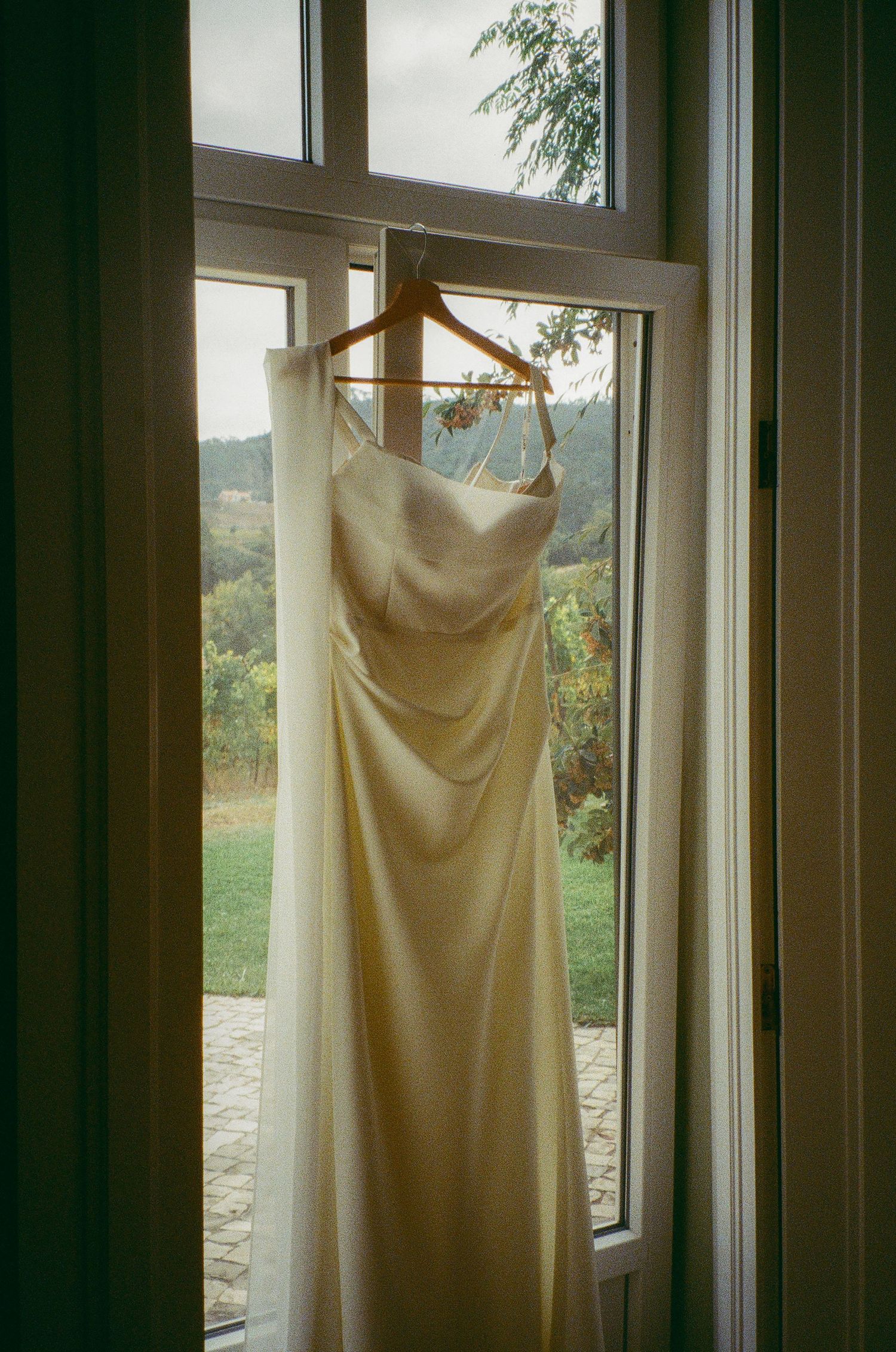Elegant ivory satin wedding dress hanging on wooden hanger in window with vineyard views.