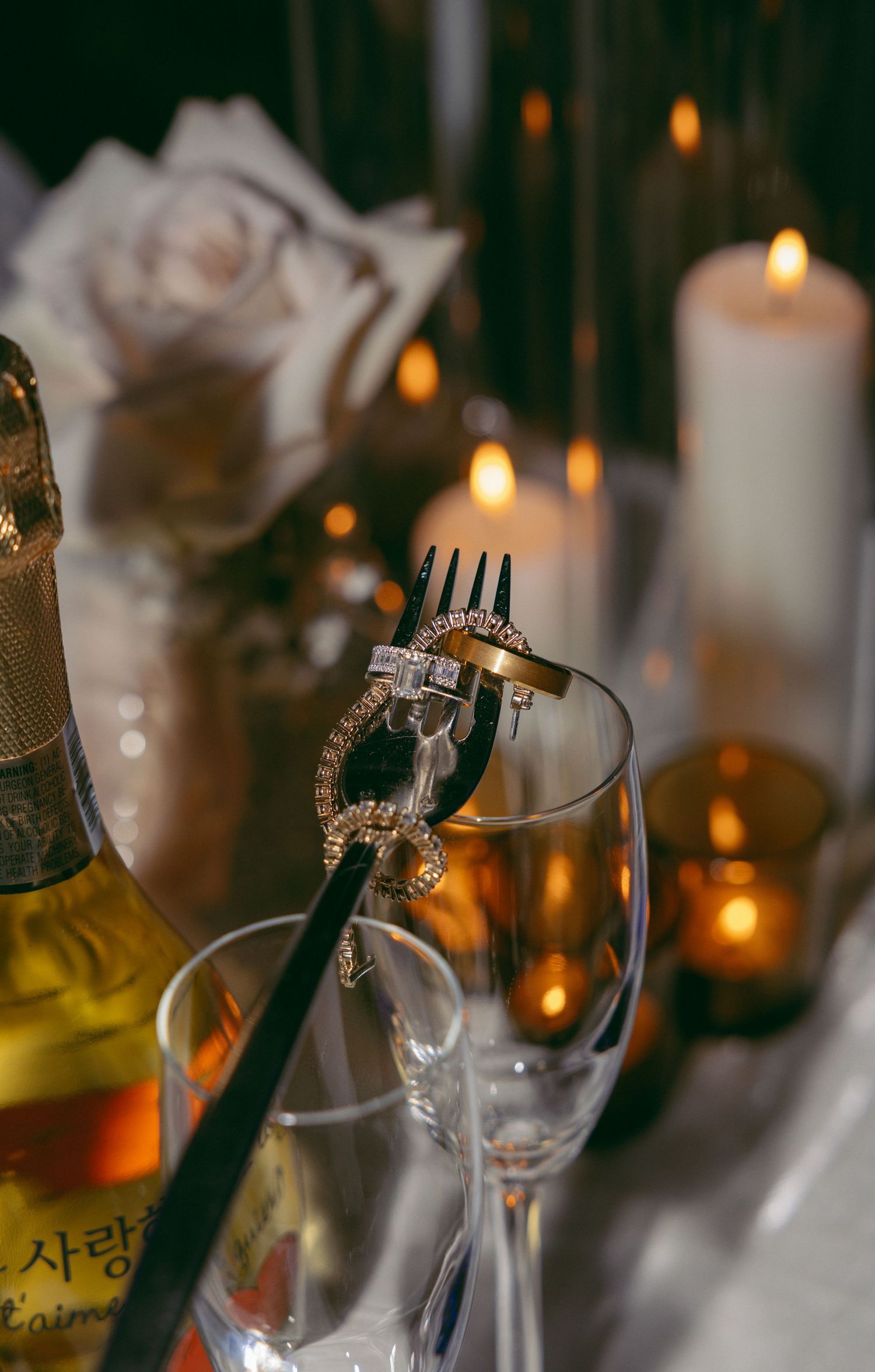 Diamond ring on a fork beside champagne glasses, candles, and a rose in a romantic engagement proposal setting.