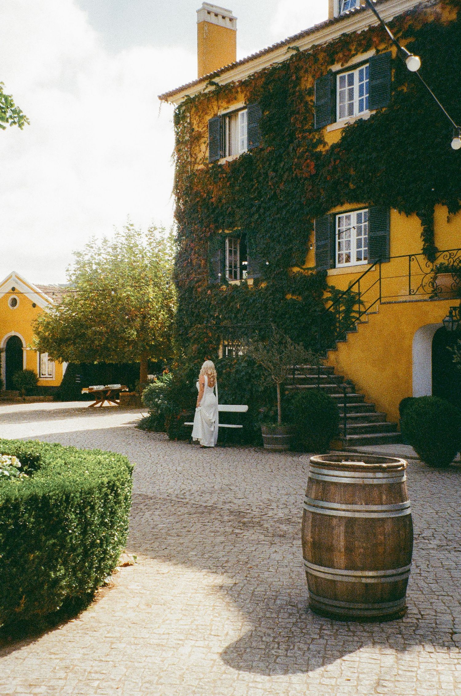 Bride in white dress stands near ivy-covered yellow estate with wooden barrel and cobblestone courtyard.