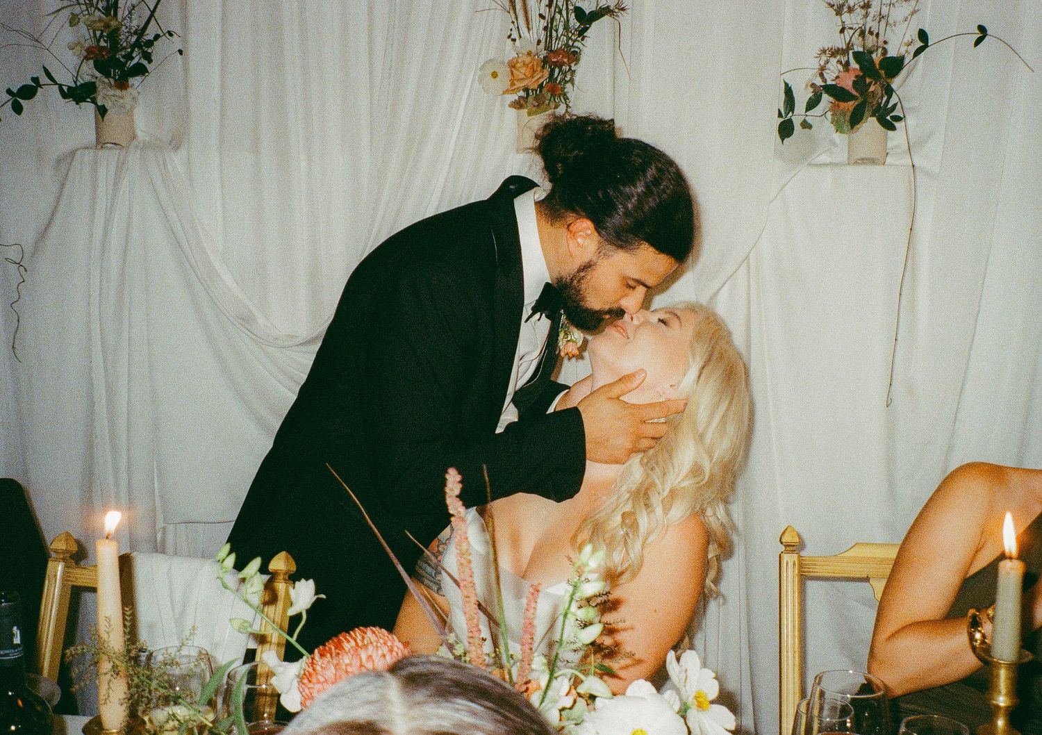 Couple sharing a romantic kiss at wedding reception, surrounded by floral arrangements and candlelight.